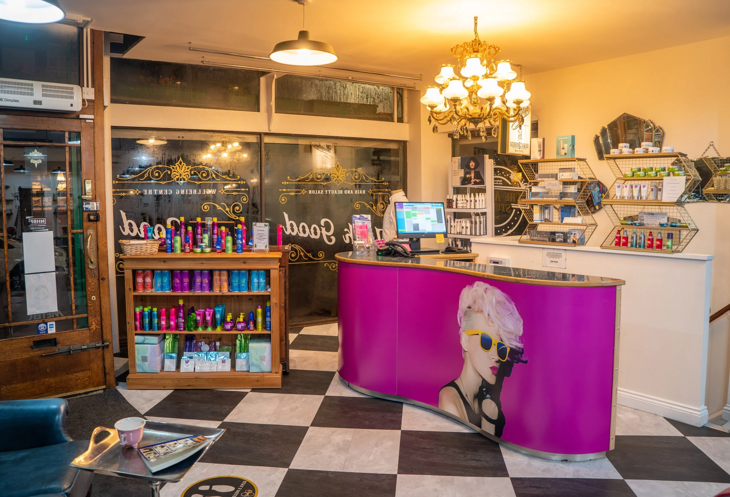 Interior of a beauty salon reception area with a curved purple counter featuring an image of a woman with blonde hair wearing sunglasses. Behind the counter, there are shelves with skincare and beauty products, a computer, and a chandelier hanging from the ceiling. To the left, there's a wooden display with colorful bottles, and the entrance door is visible with gold decorative text on the glass.
