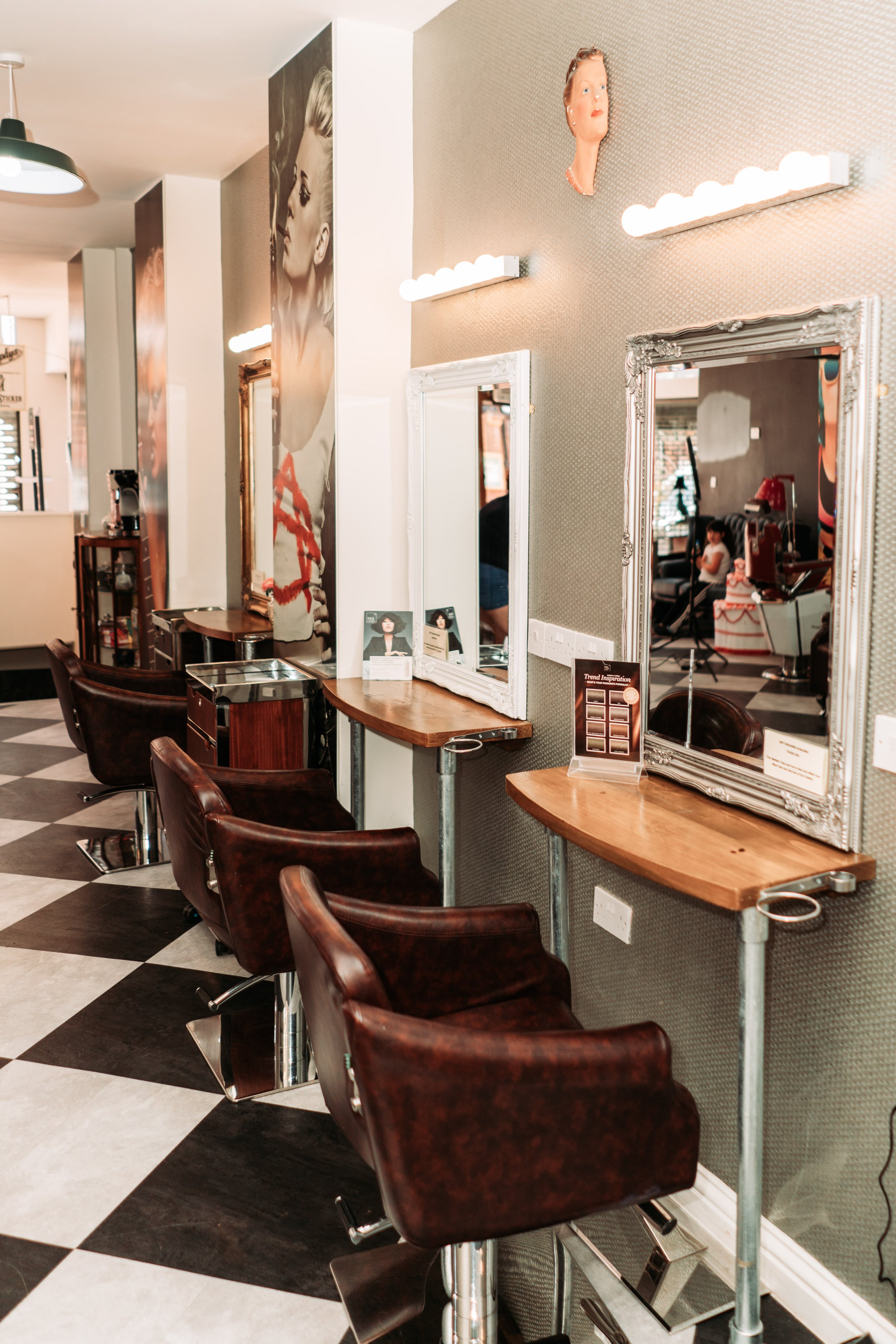 Interior of a hair salon with four styling chairs, mirrors, and wooden counters. There are artworks and photographs on the wall, and the salon has a checkered black and white floor.