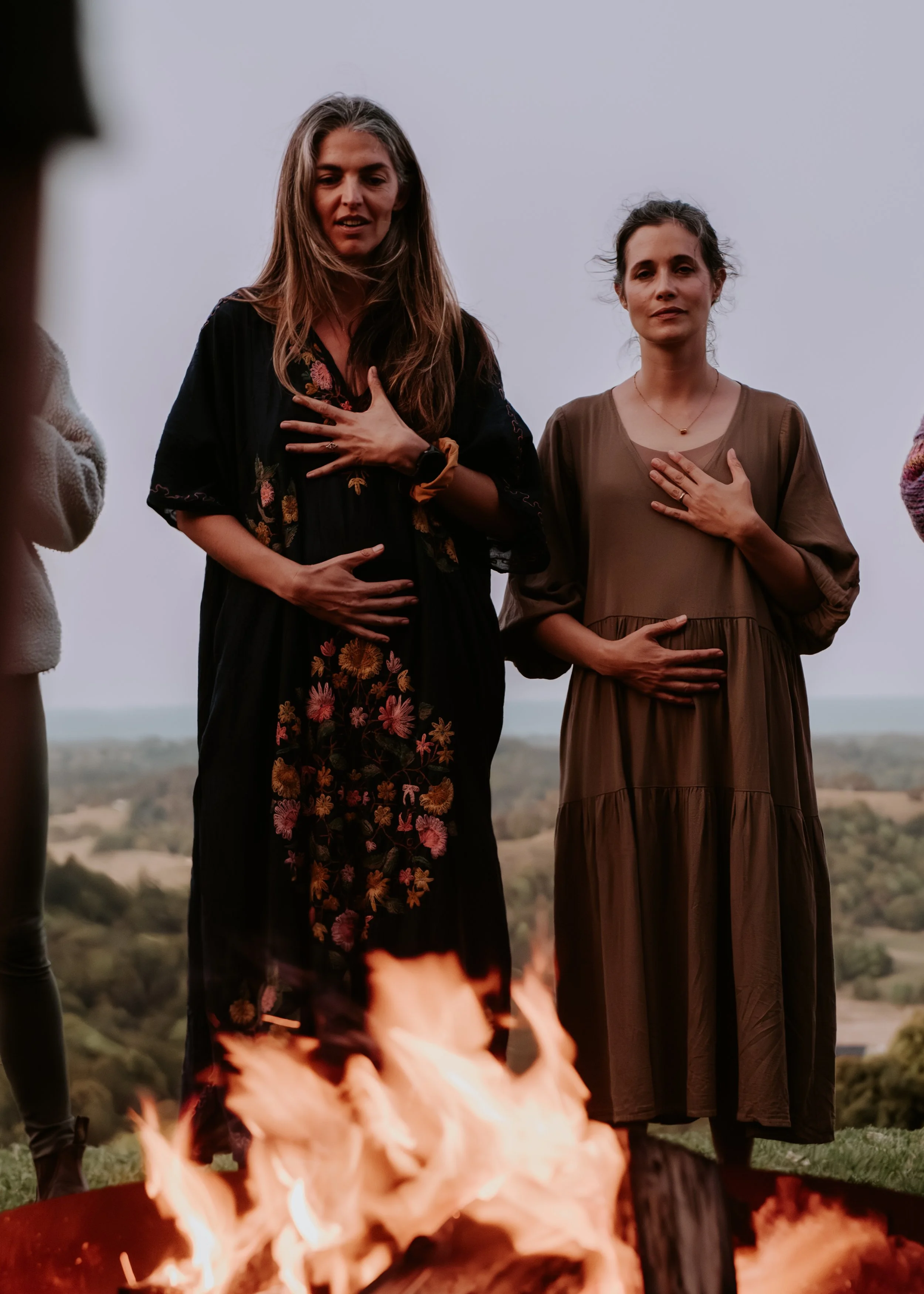 Two women standing outdoors with their hands over their chests, near a campfire, under a cloudy sky.