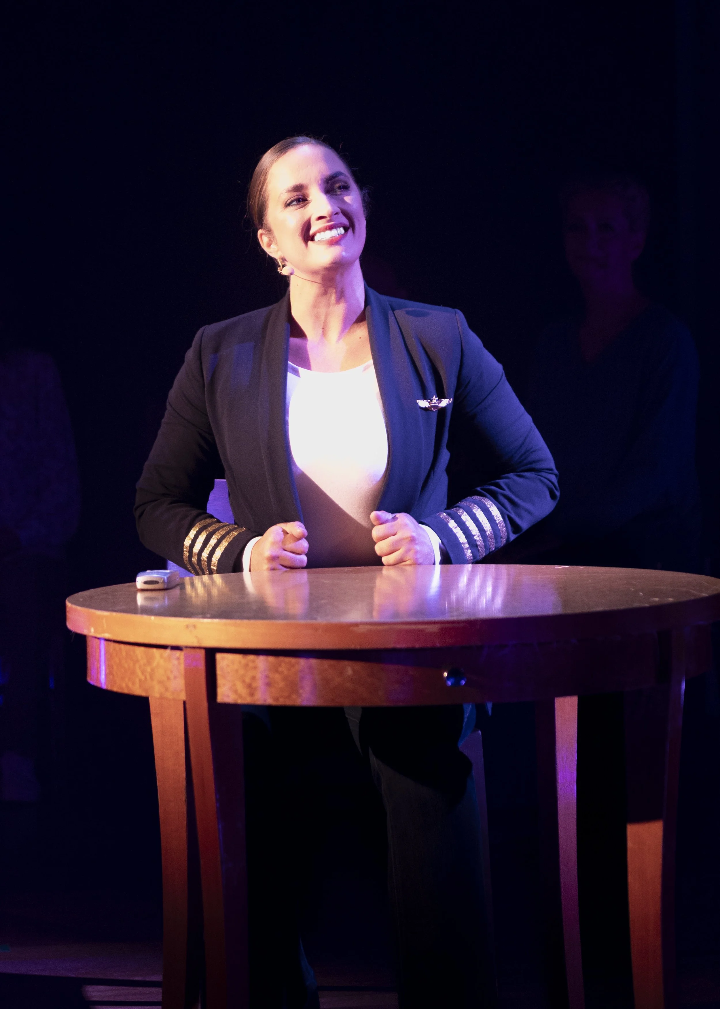A woman in a navy blazer with gold-striped cuffs and a white top is smiling and standing behind a wooden table under stage lighting.