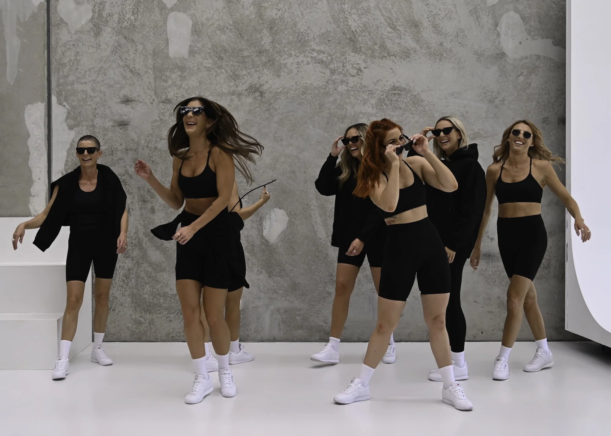 Six women dancing and smiling, wearing black athletic outfits and sunglasses, in a bright indoor space with a gray textured wall background.