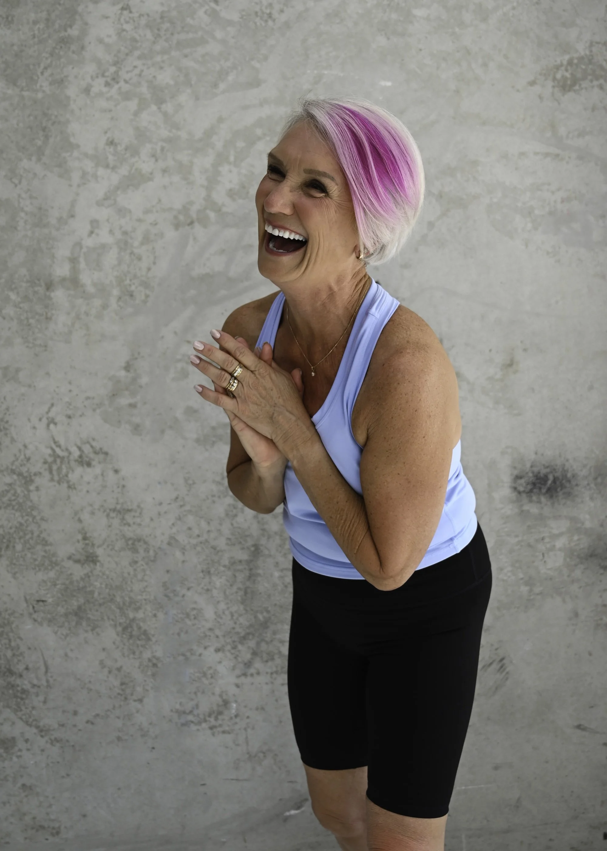 An older woman with short, pink-tinted hair, smiling widely and holding her hands together near her chest, standing against a textured gray wall. She is wearing a light blue tennis tank top and black shorts.