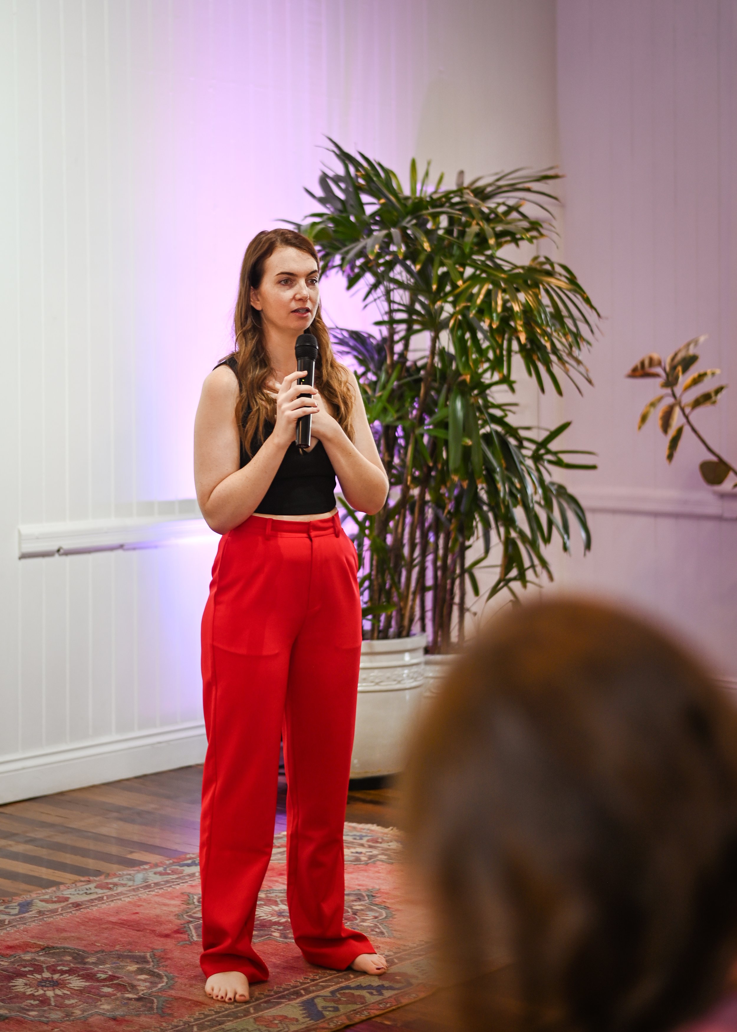 A woman with long brown hair, wearing a black sleeveless top, and bright red pants, is speaking into a microphone in a room with a large green plant and purple lighting.