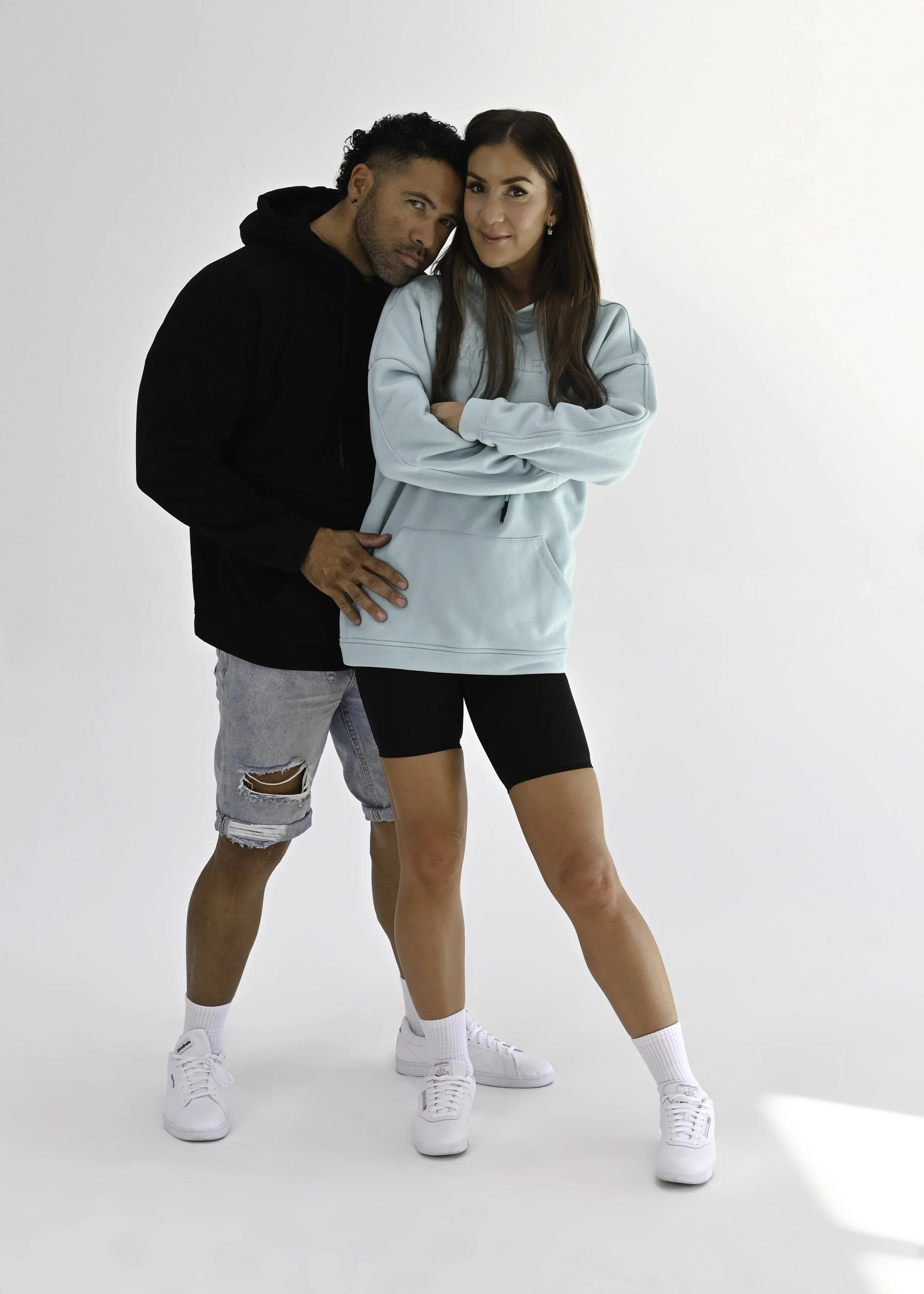 A man and a woman standing together against a white background. The man is wearing a black hoodie, distressed denim shorts, and white sneakers. The woman is wearing a light blue hoodie, black shorts, and white sneakers, with her arms crossed. They ar