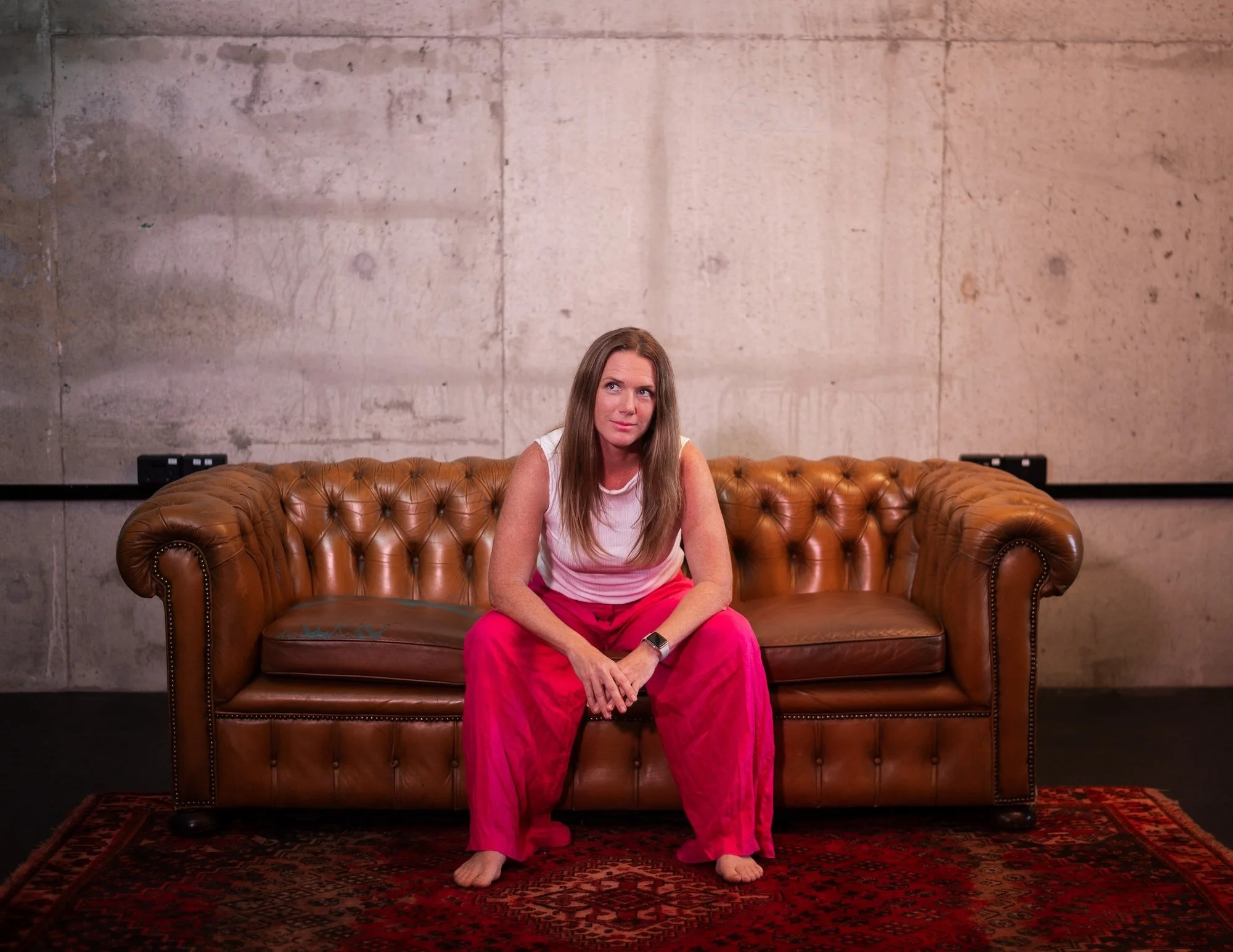 A woman with long brown hair sitting on a brown leather tufted sofa in a room with concrete walls and a rug in front of her.
