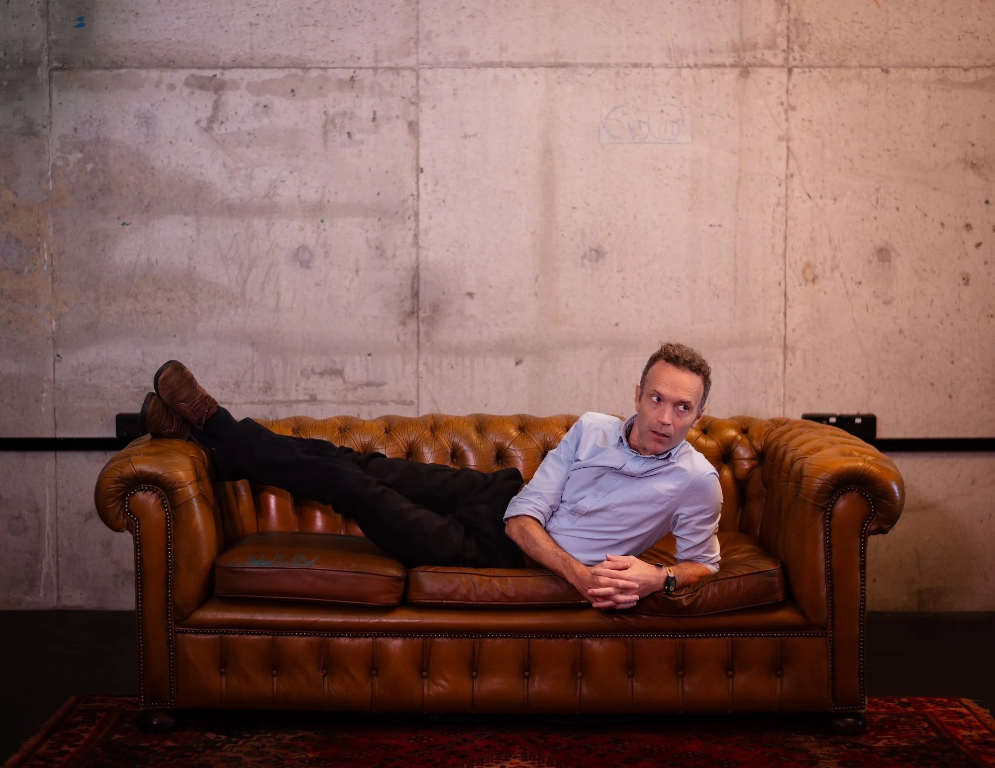 A man lying on a brown leather couch in front of a concrete wall, looking to his right.