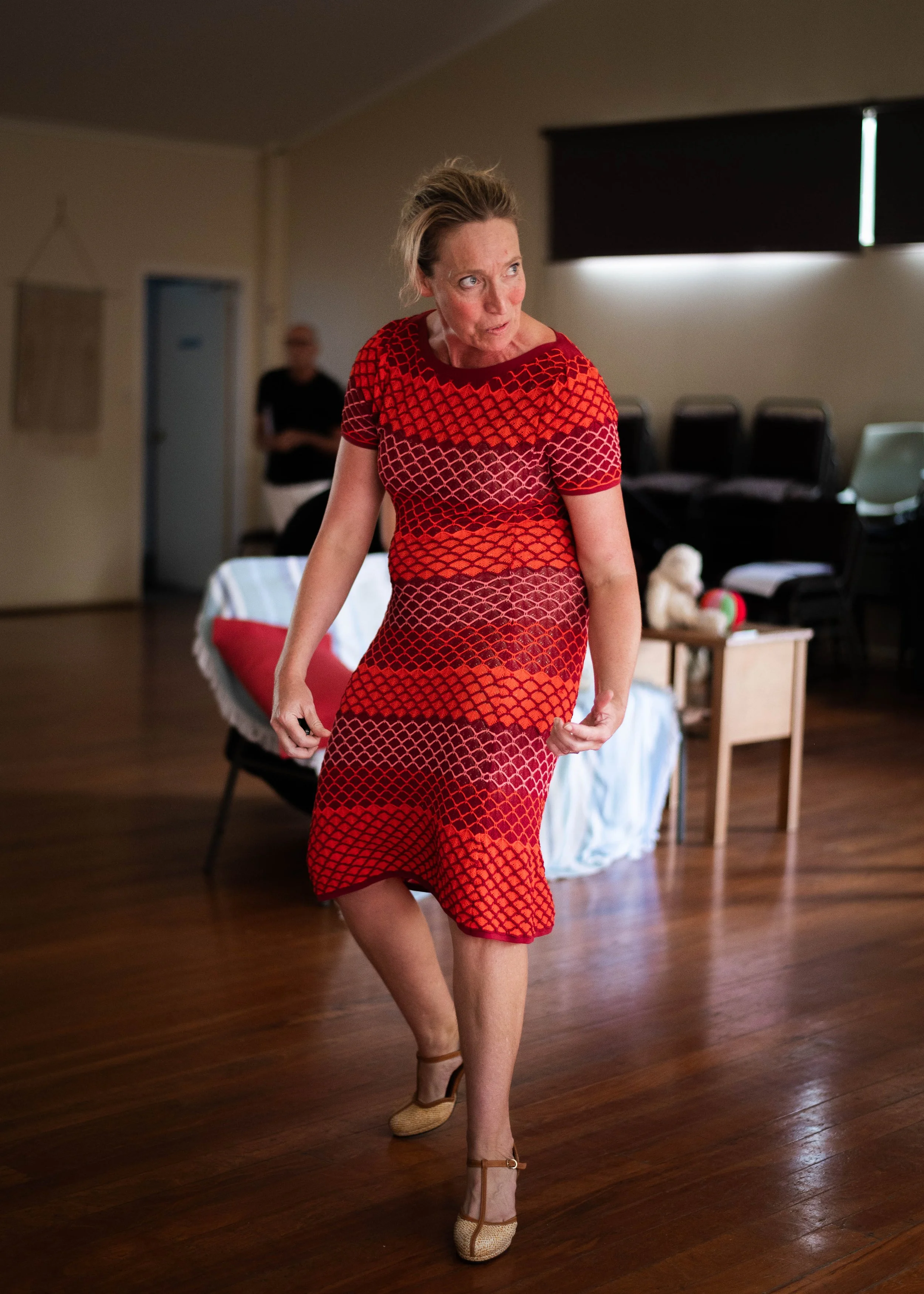 A woman in a red and black patterned dress dancing indoors, with a person blurred in the background and a dog toy on a table nearby.