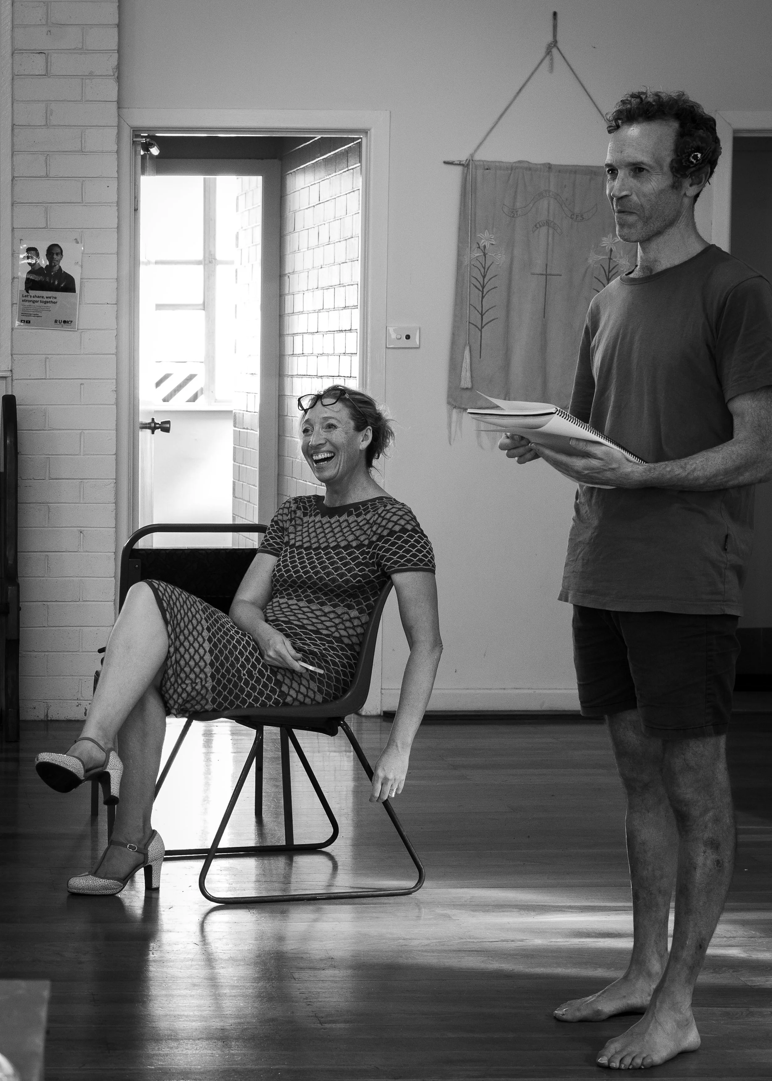 A woman in a patterned dress and high heels laughs while sitting on a chair, and a man in casual clothing and barefoot stands nearby holding a notebook, in an indoor room with a door, window, and wall hanging.