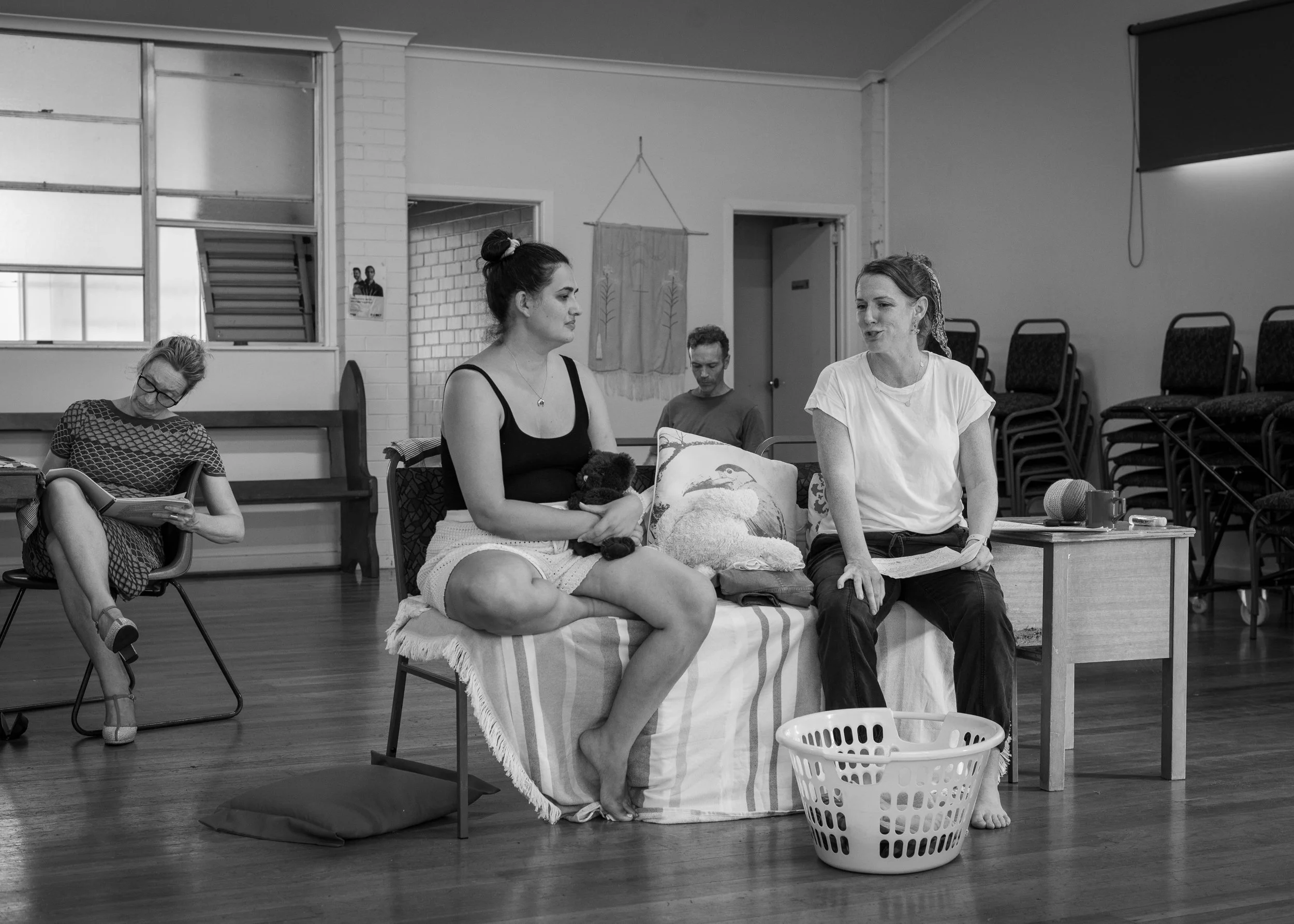A group of five people in a room with stacked chairs, engaged in discussion. Two women are sitting on a sofa and a chair, one with a teddy bear, talking. The others are seated apart; one reading a book, and one looking down.
