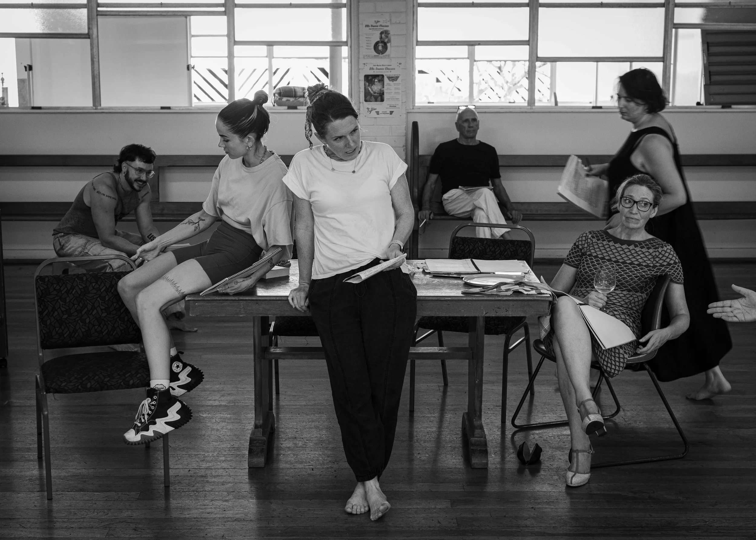 Rehearsal or meeting with six people in a dance studio or rehearsal space, some sitting, some standing, with a window and posters on the wall in the background.