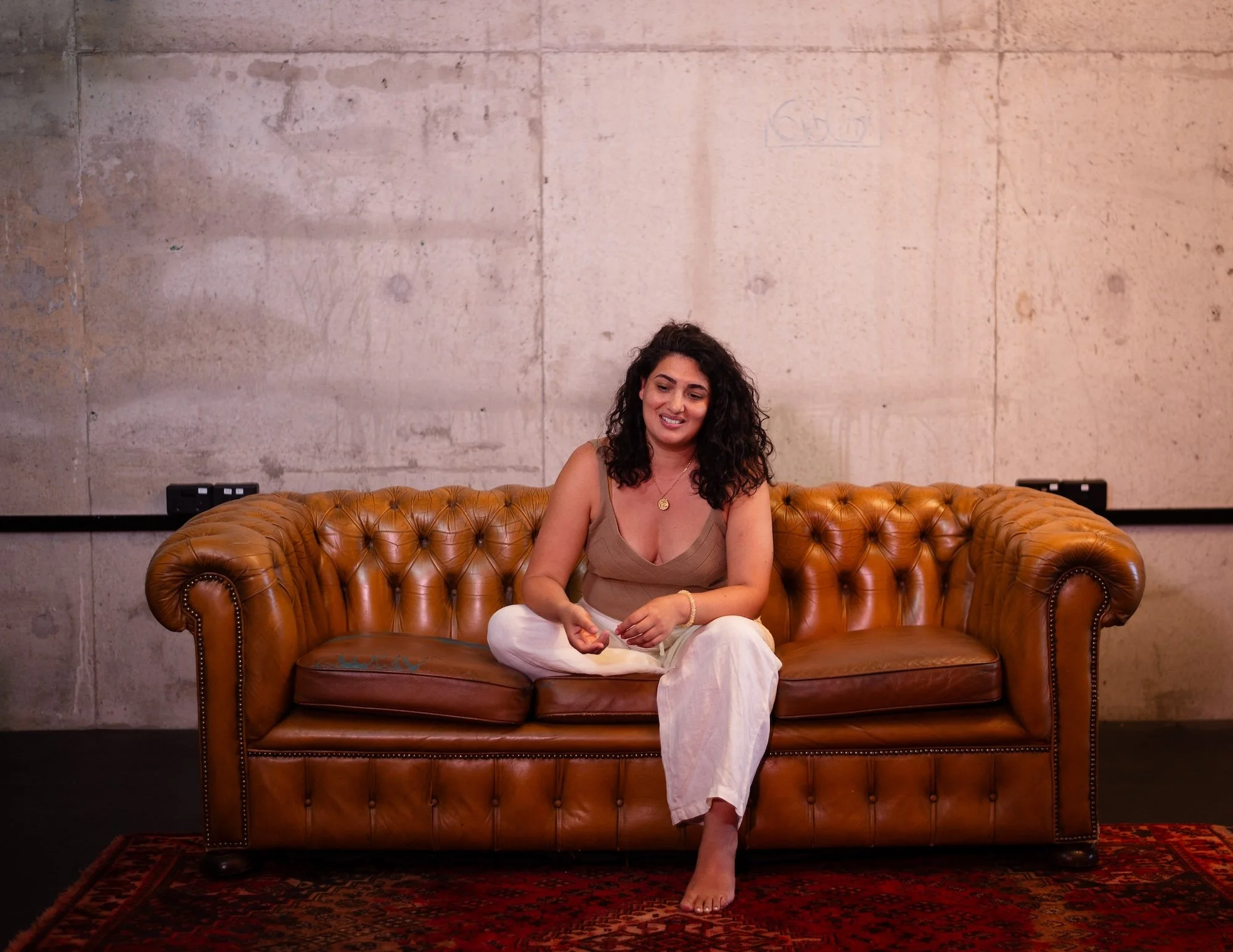 A woman with curly dark hair sitting on a brown leather couch in front of an unfinished concrete wall.