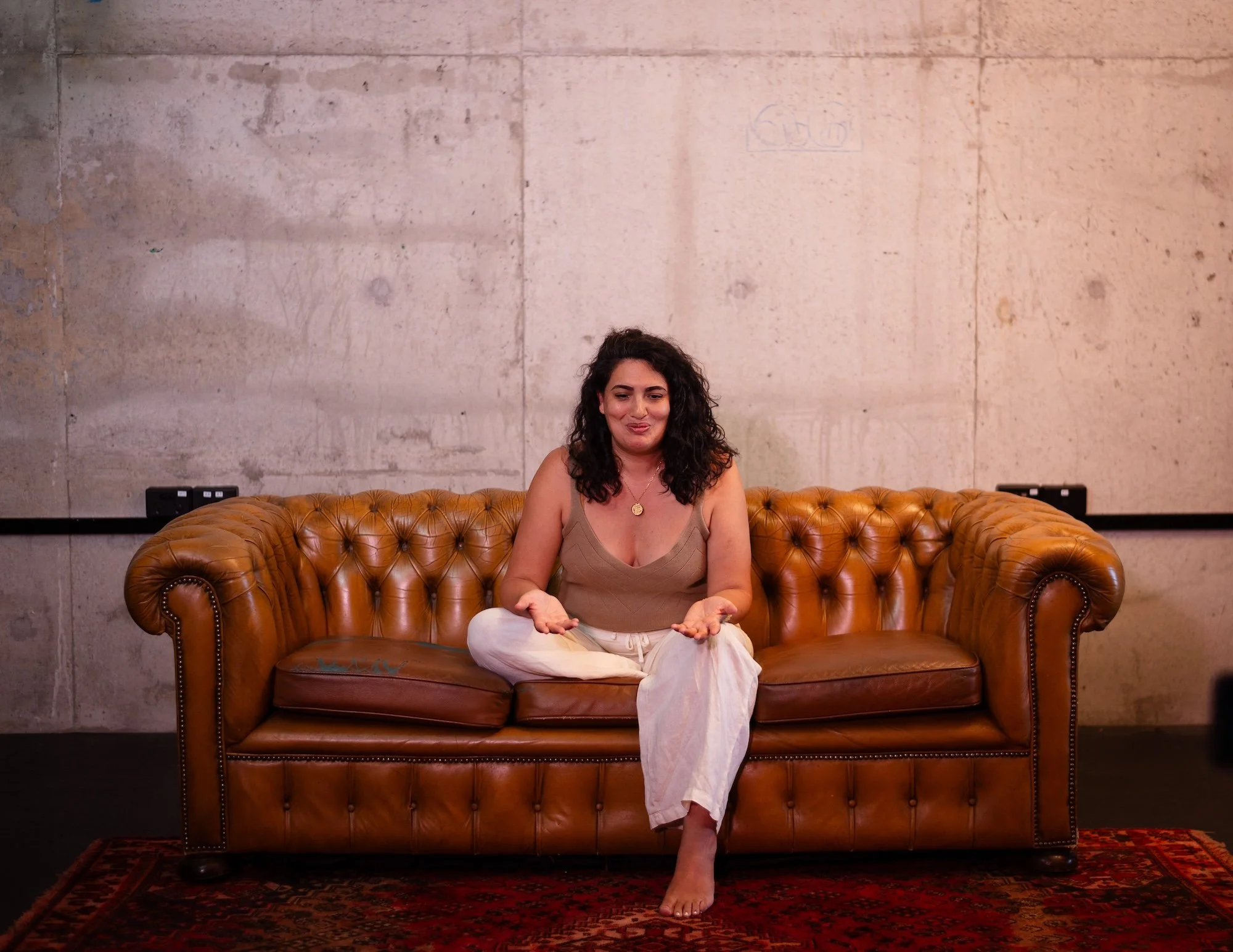 A woman with curly dark hair sits cross-legged on a brown leather tufted sofa, gesturing with open hands, against a plain concrete wall.