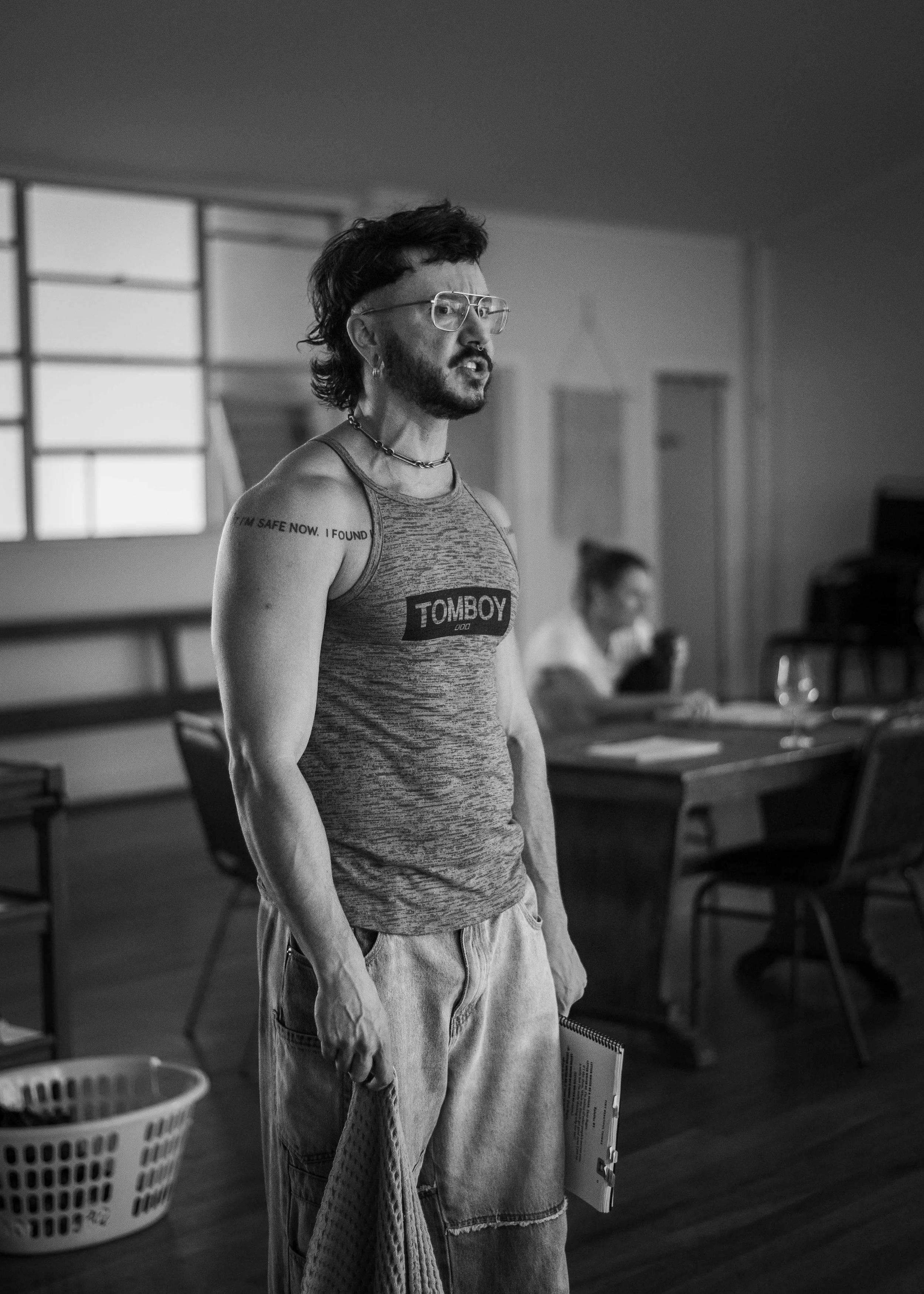 Black and white photo of a man with a beard, glasses, and a mohawk hairstyle, wearing a sleeveless shirt with "TOMBOY" printed on it, standing in a room with tables and chairs, holding a notebook or spiral-bound pad in his right hand.