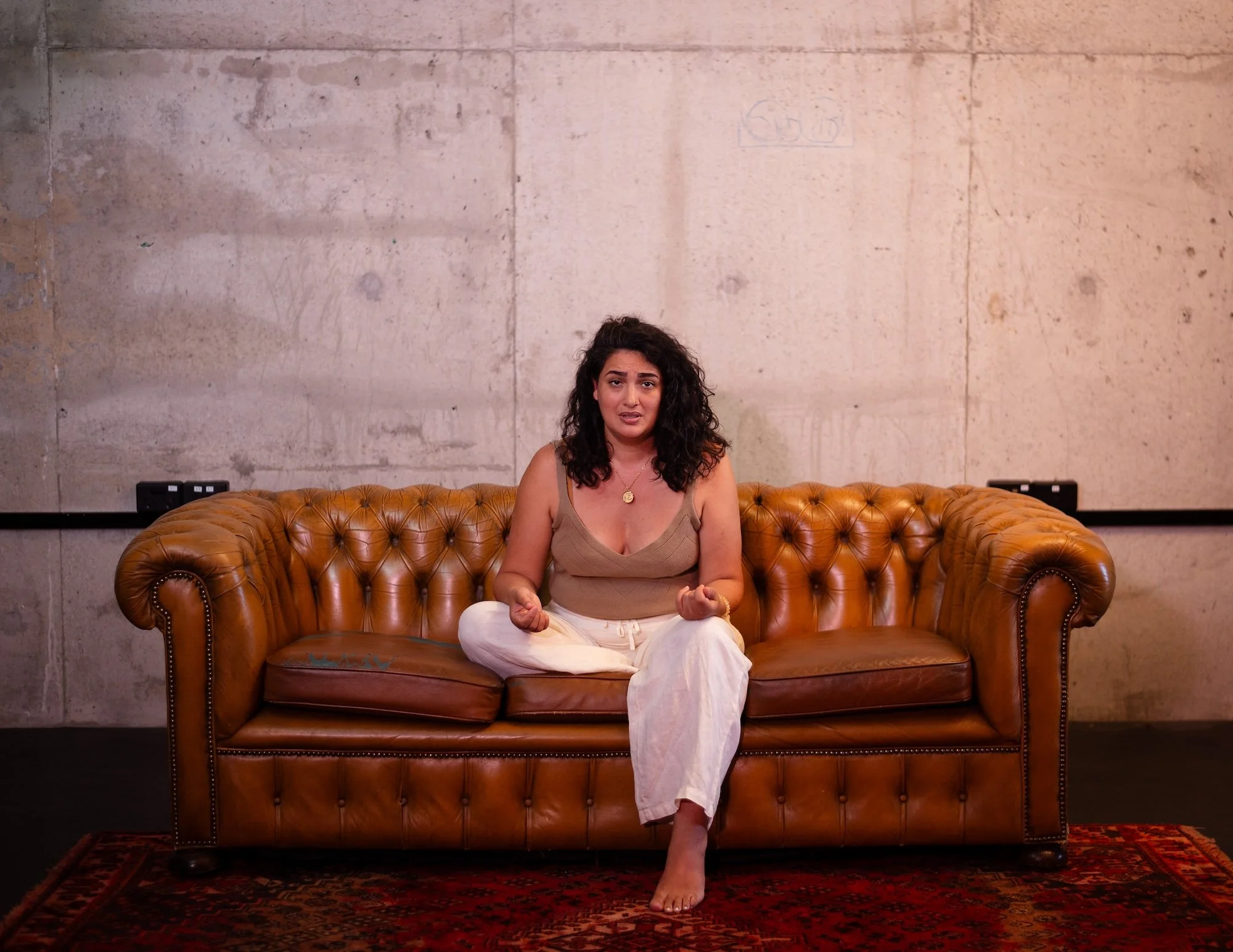 A woman with curly dark hair sitting cross-legged on a brown leather couch against a concrete wall.