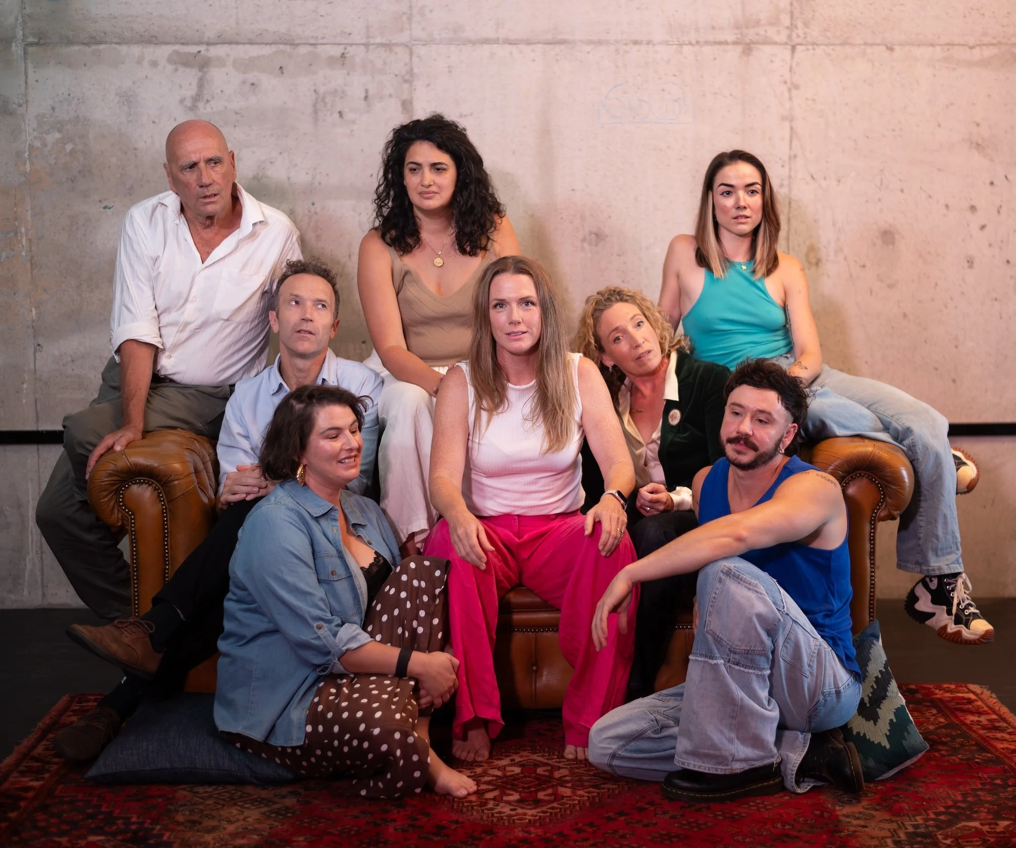 A group of nine diverse people posing together indoors against a plain wall, some sitting on a leather couch and others sitting or kneeling on a rug in front.