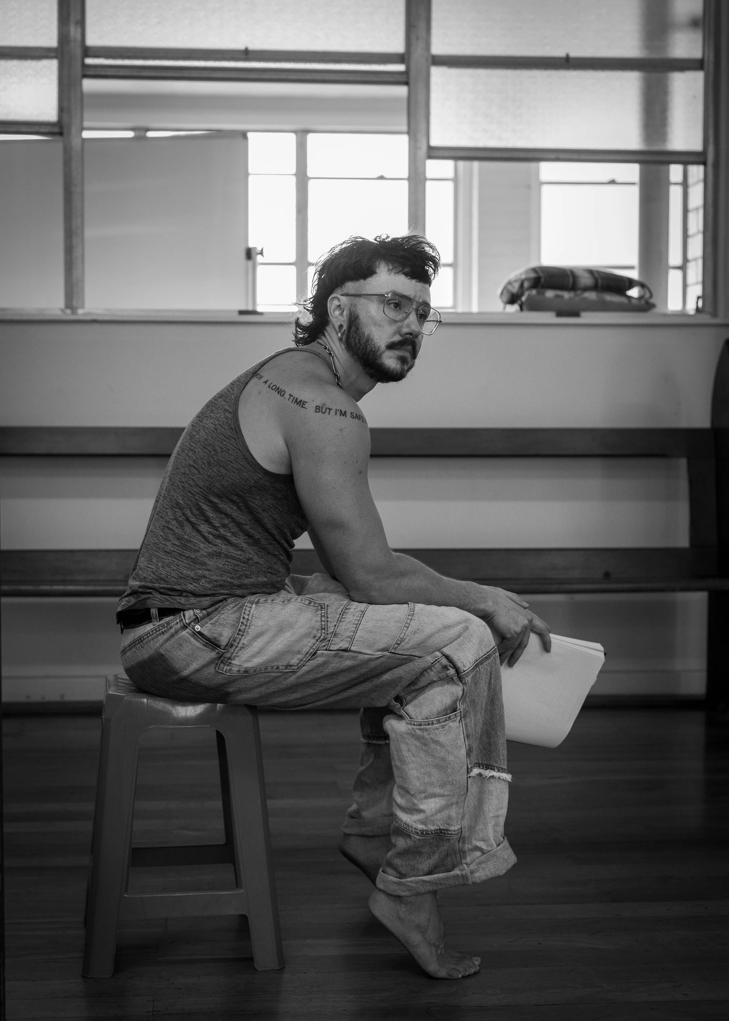 A man with glasses and a beard, sitting barefoot on a stool, holding a notebook, in a room with large windows and wooden floors, in black and white.