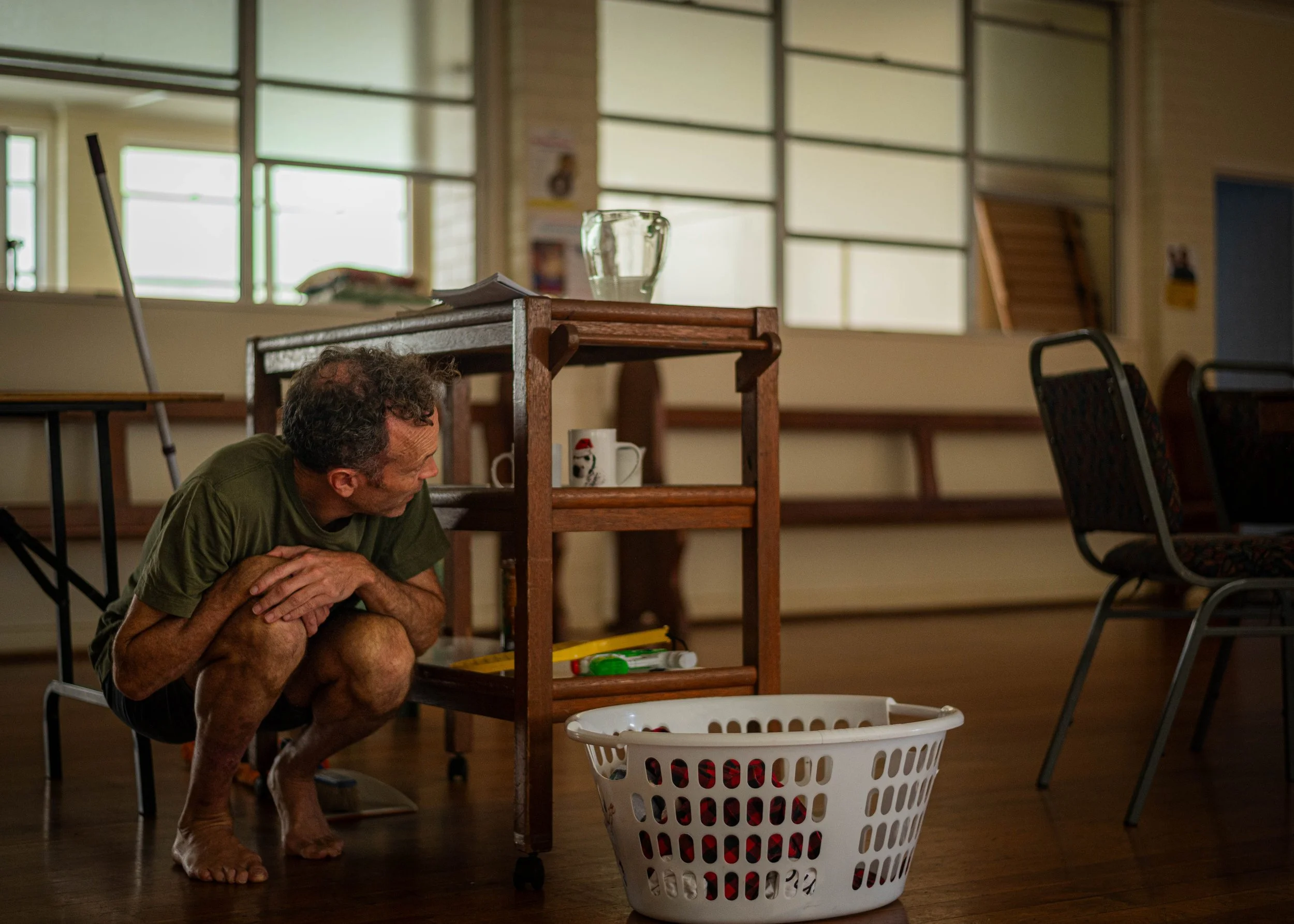 A man crouching behind a wooden cart in a room with large windows, a white laundry basket in front, and several chairs and items on the cart.