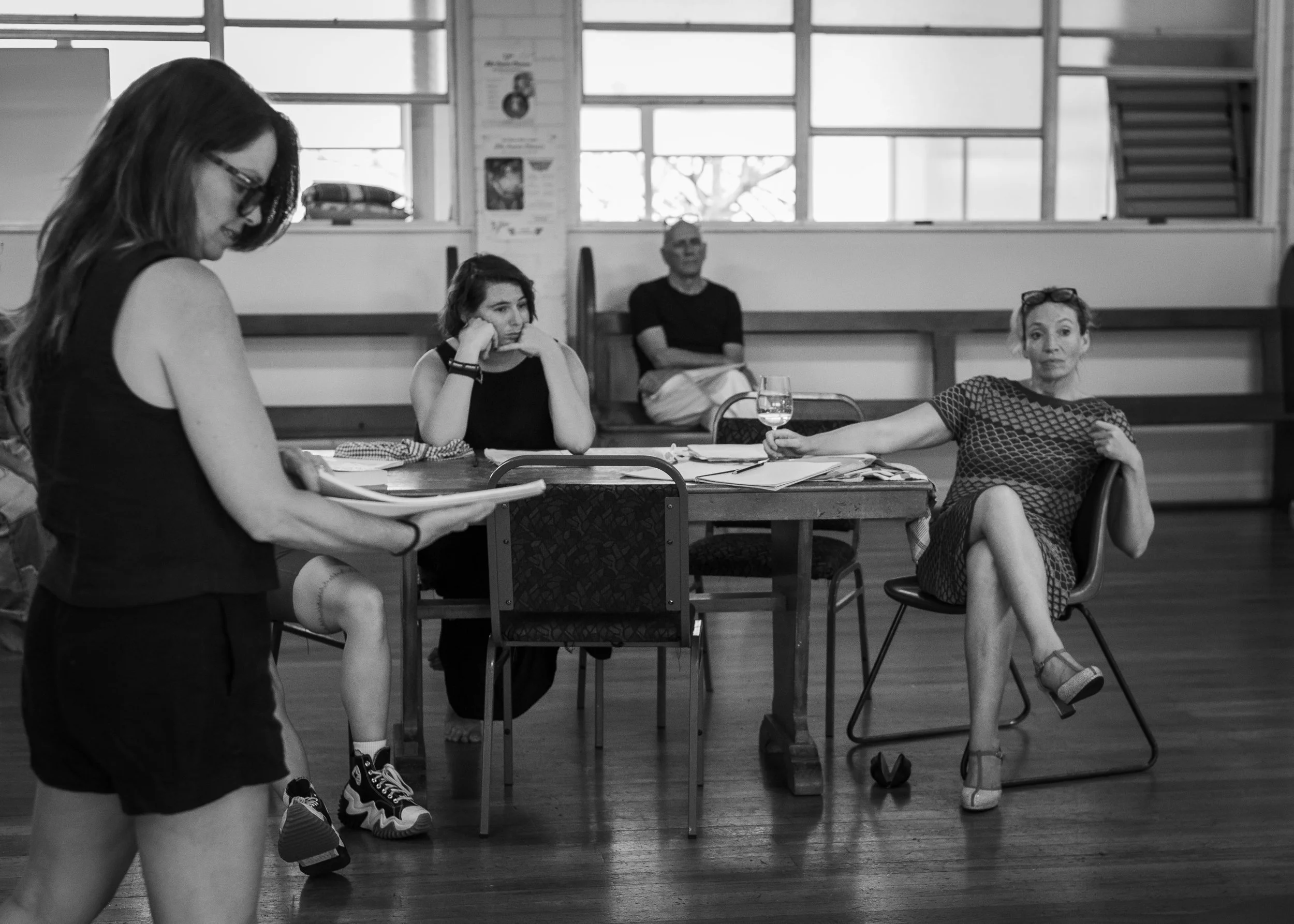 A woman is standing and reading or reviewing notes, while three other people are seated at a table. The woman on the right holds a glass of water, and there are papers and a notebook on the table. The scene appears to be in a rehearsal or meeting spa