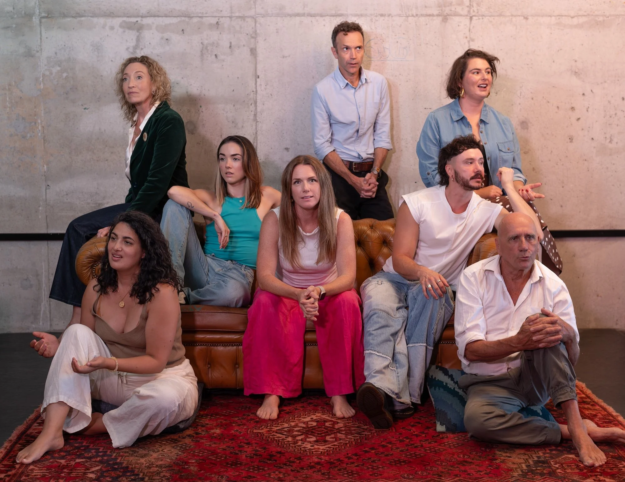 Group of nine diverse people sitting and standing on a vintage couch and floor in an industrial-style room, appearing to watch a performance or presentation.