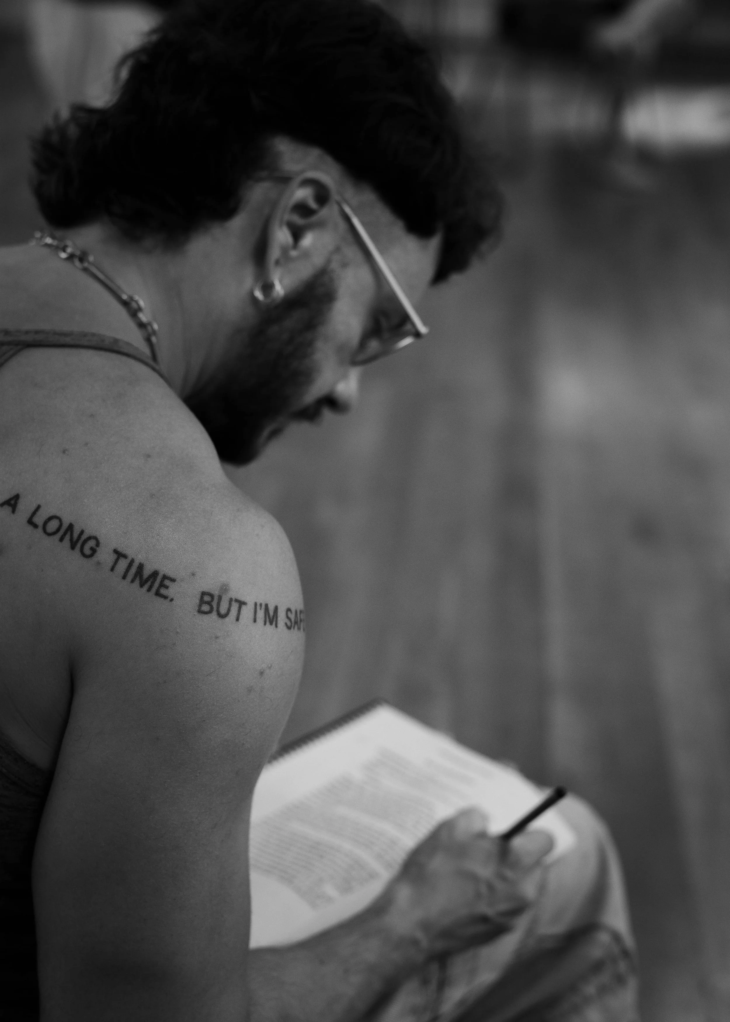 A man with glasses, a beard, and earrings is sitting and reading a book, holding a pencil, with a tattoo on his shoulder that reads 'A LONG TIME, BUT I'M SAFE.'