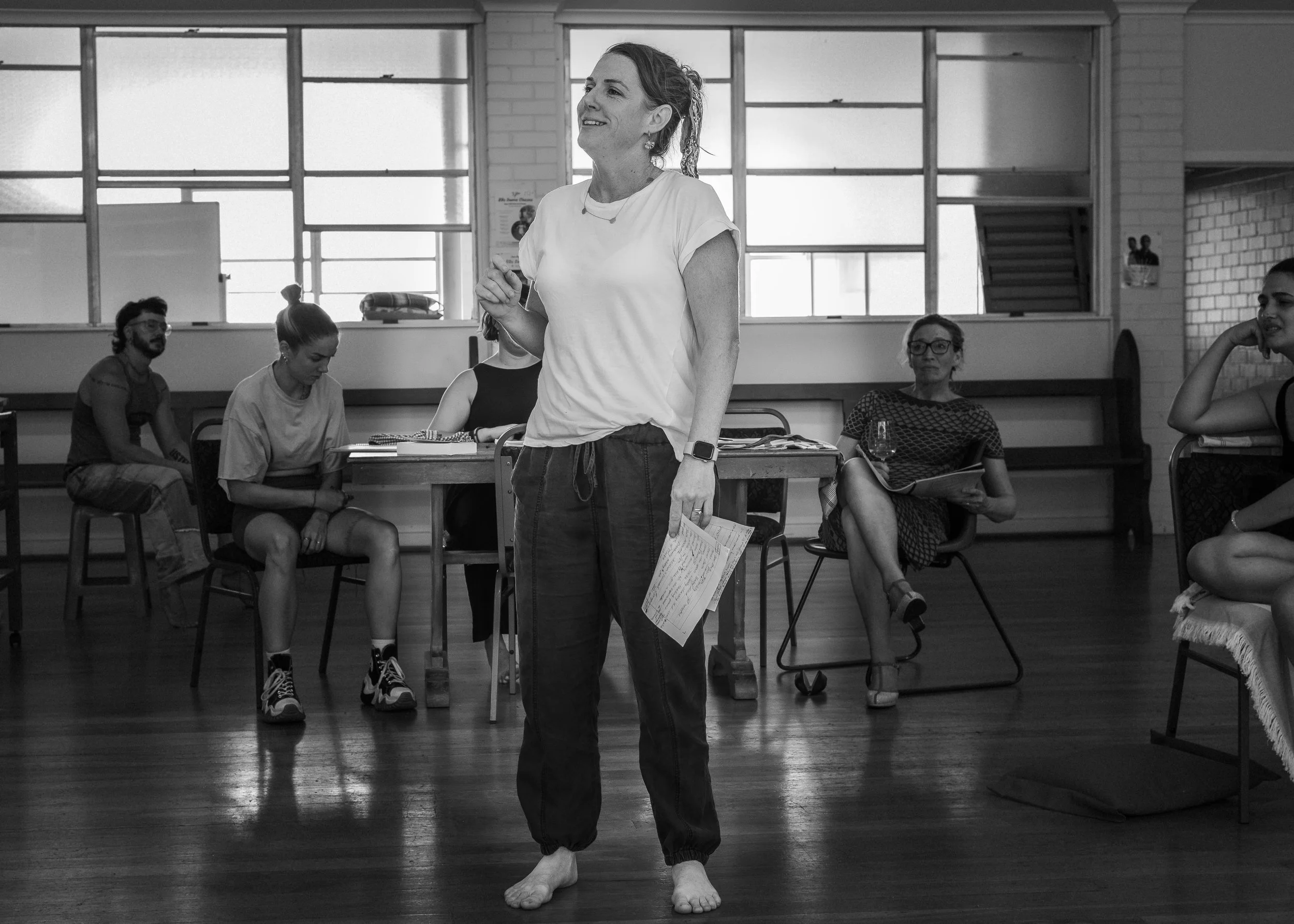 A woman standing barefoot in a dance studio, holding papers, with a group of women seated behind her, some reading and others listening.