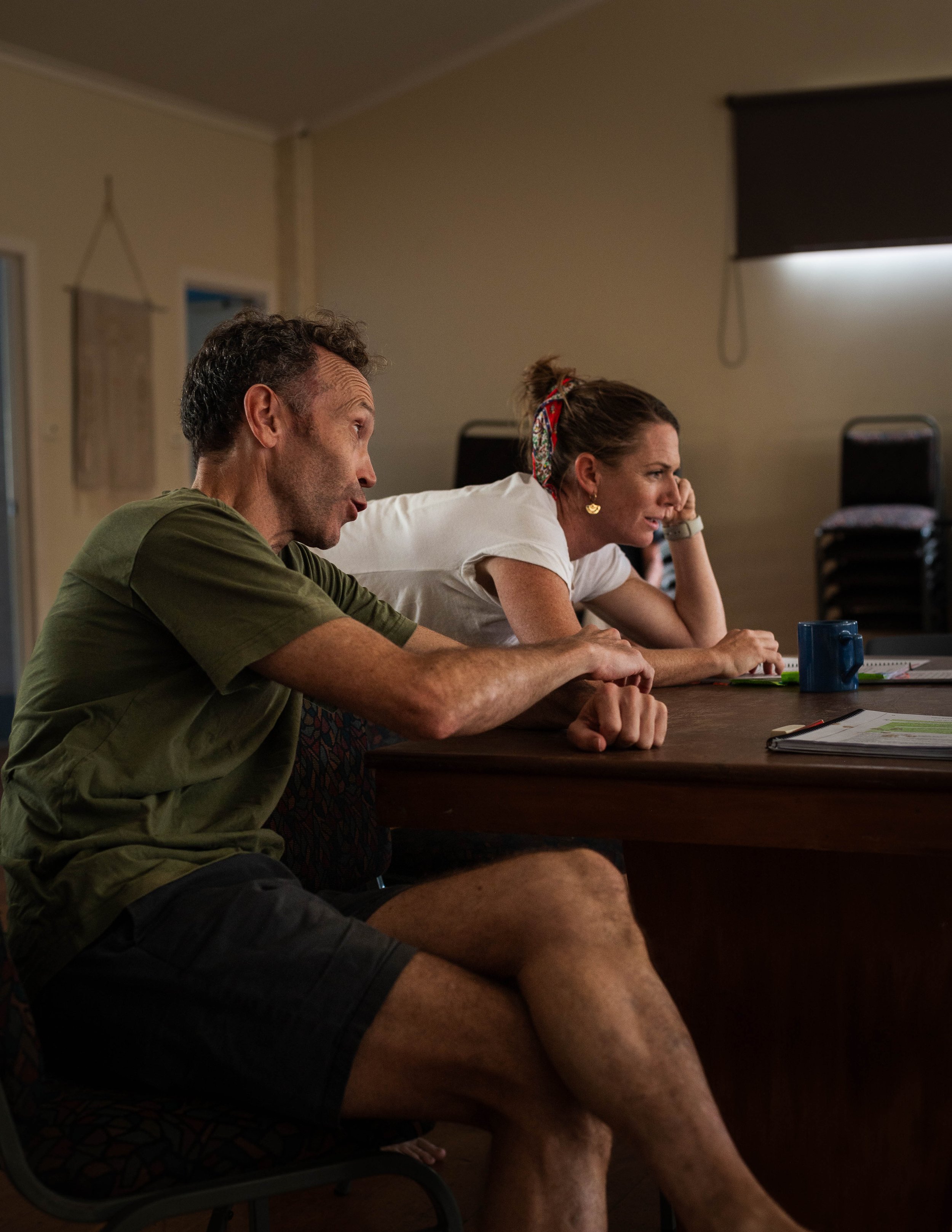 Two people sitting at a table in a dimly lit room, engaged in conversation, with a stack of chairs and window shades in the background.