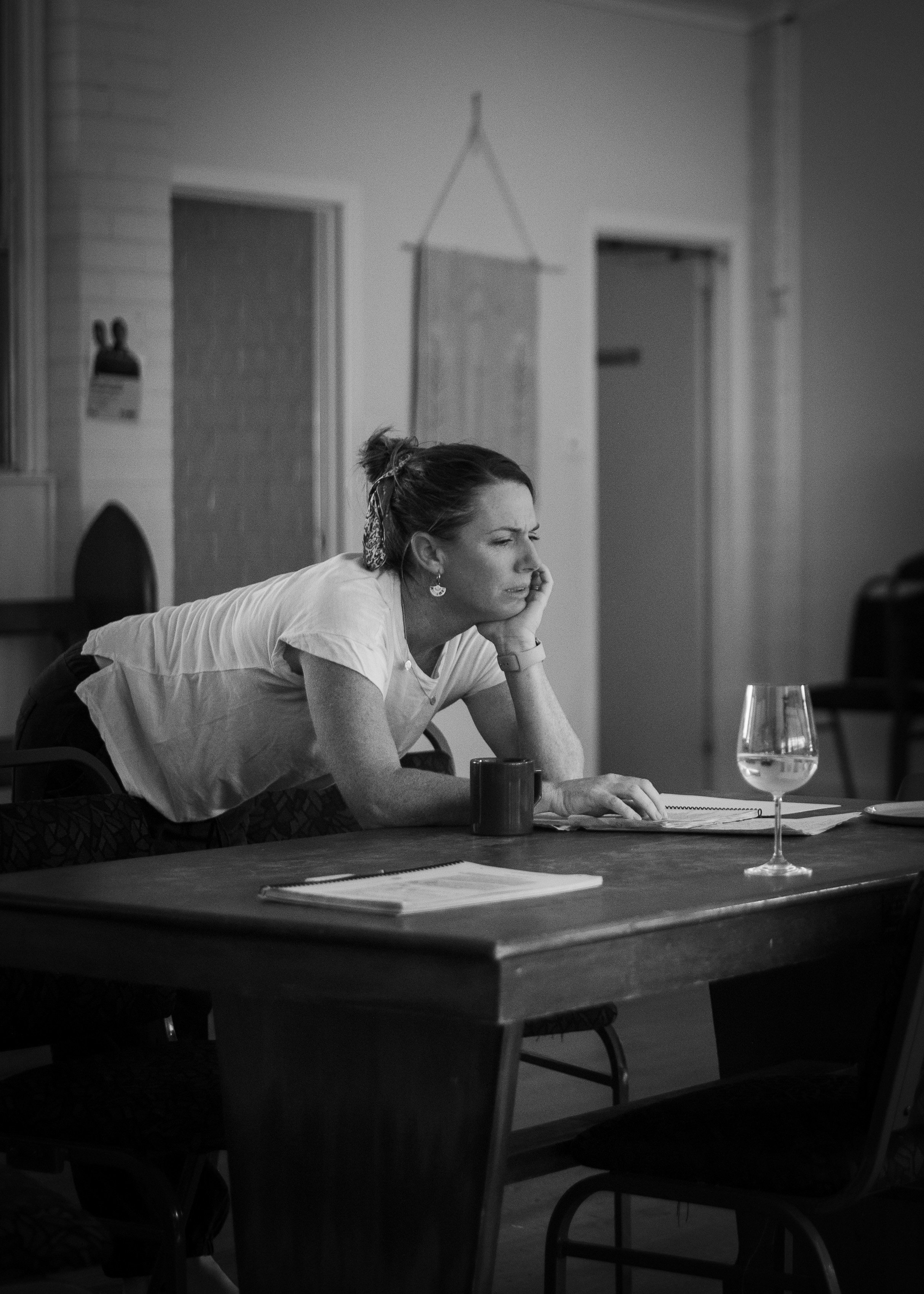 A woman with a scrunchie in her hair leans on a table, looking thoughtful, with a glass of water, a mug, and some papers in front of her.