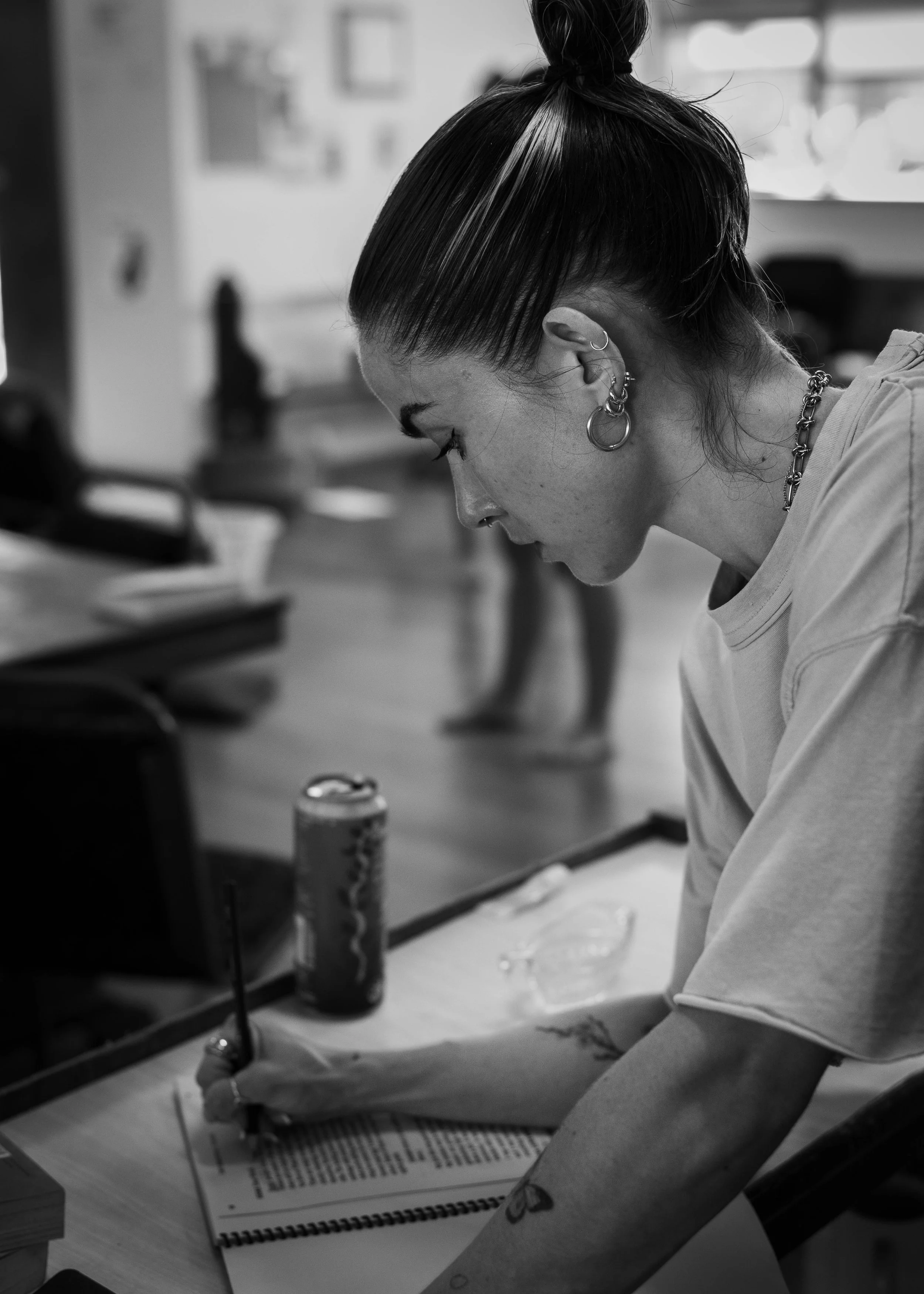 A woman with multiple earrings and a tattoo on her arm is writing on a piece of paper at a desk, with a can of drink nearby; photo in black and white.