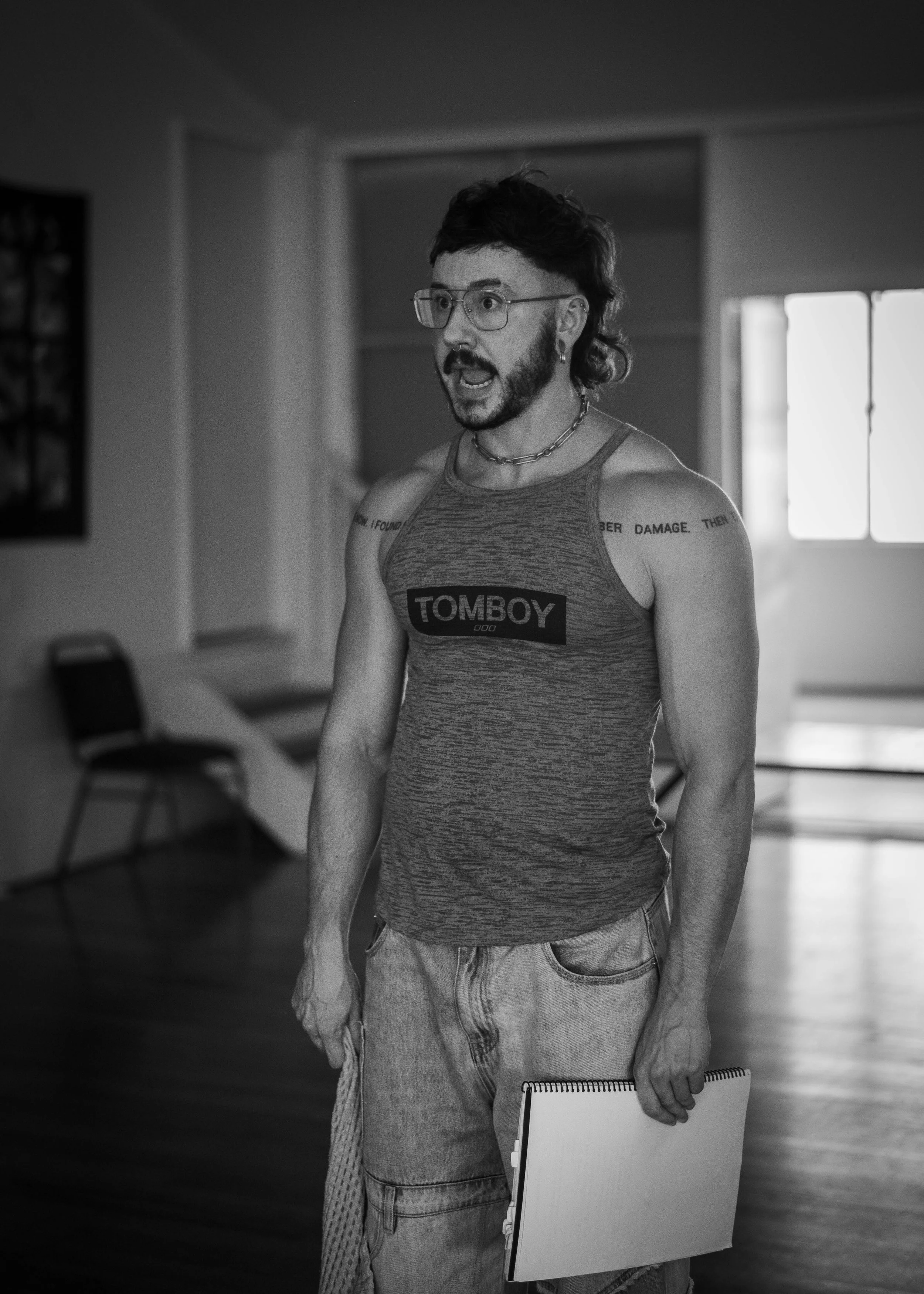A person with glasses, a beard, and tattoos, wearing a tank top and baggy pants, holding a notebook in a room with large windows and a chair, appears to be speaking.