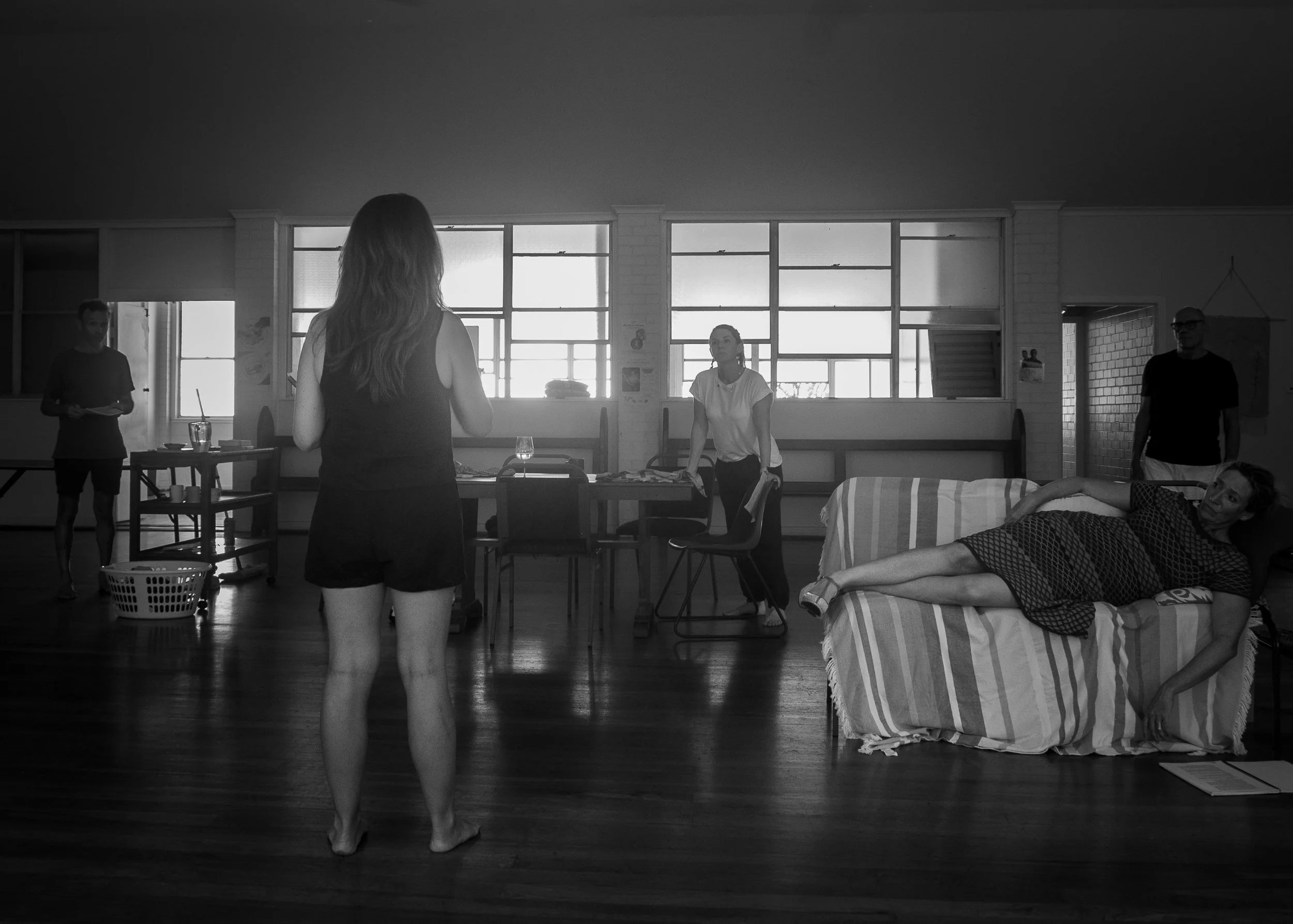 A woman standing in front of a group of people in a room with large windows, some people seated or lying on furniture, in black and white.