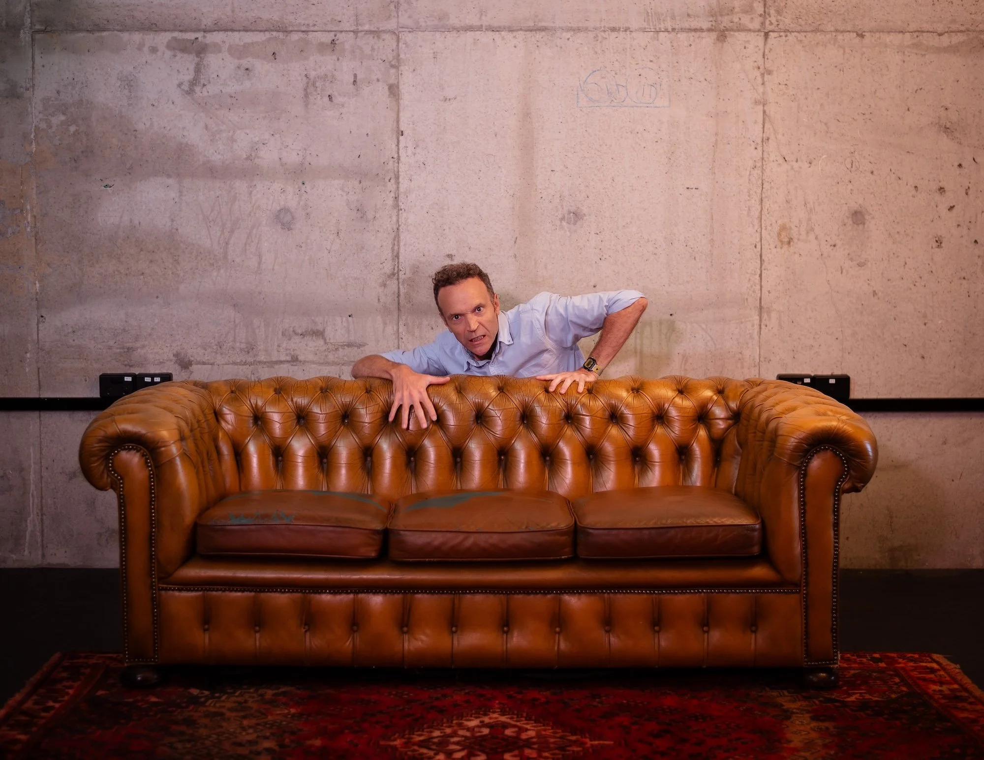 A man appears to be crawling over a vintage brown leather tufted sofa with a distressed look, set against a plain, raw concrete wall.
