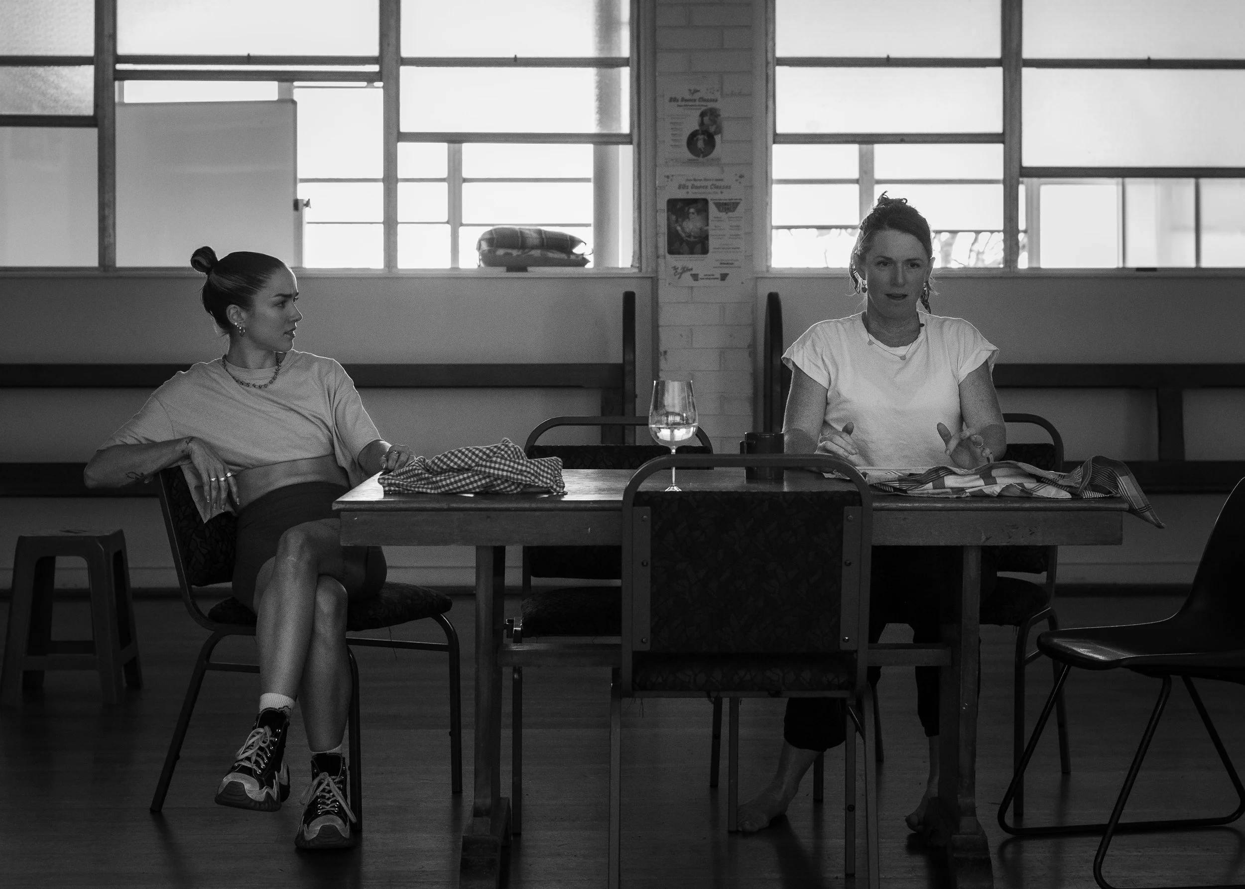 A black and white photo of two women sitting at a table inside a room with large windows. The woman on the left has her hair in a bun and is wearing a t-shirt and shorts, sitting with her legs crossed. The woman on the right has her hair tied back an