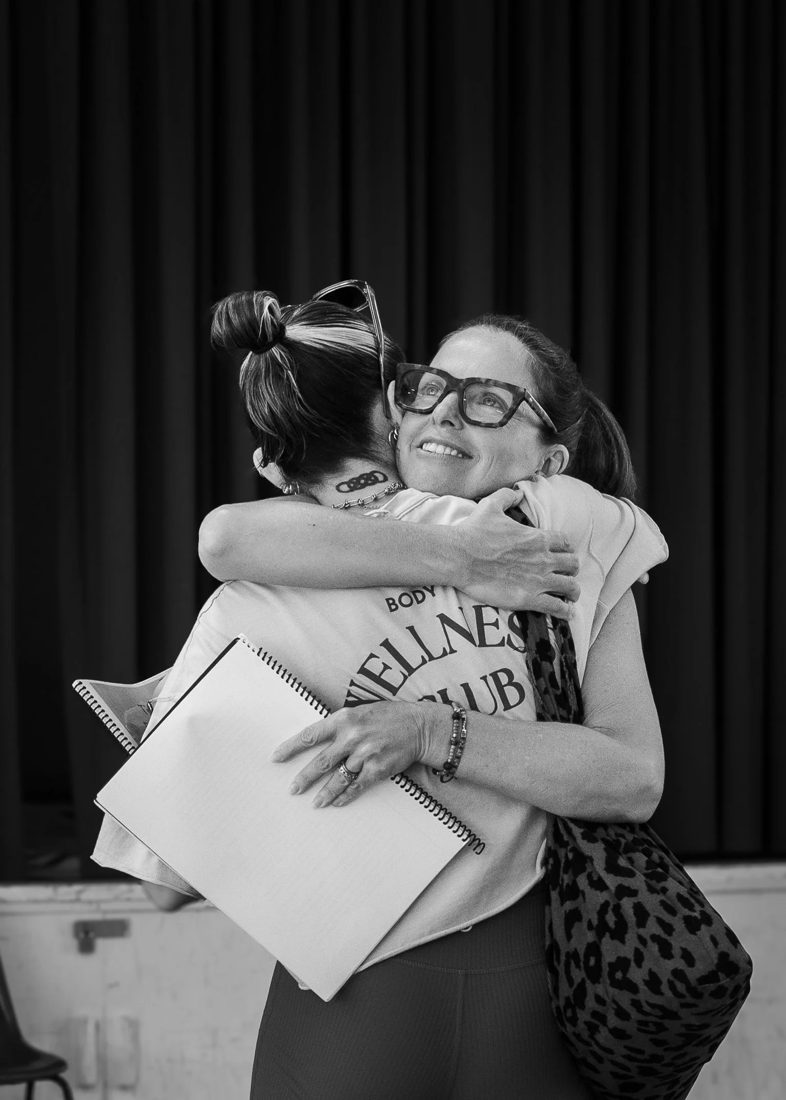 Two women hugging warmly, one with glasses and the other with a headscarf, in front of a curtain.
