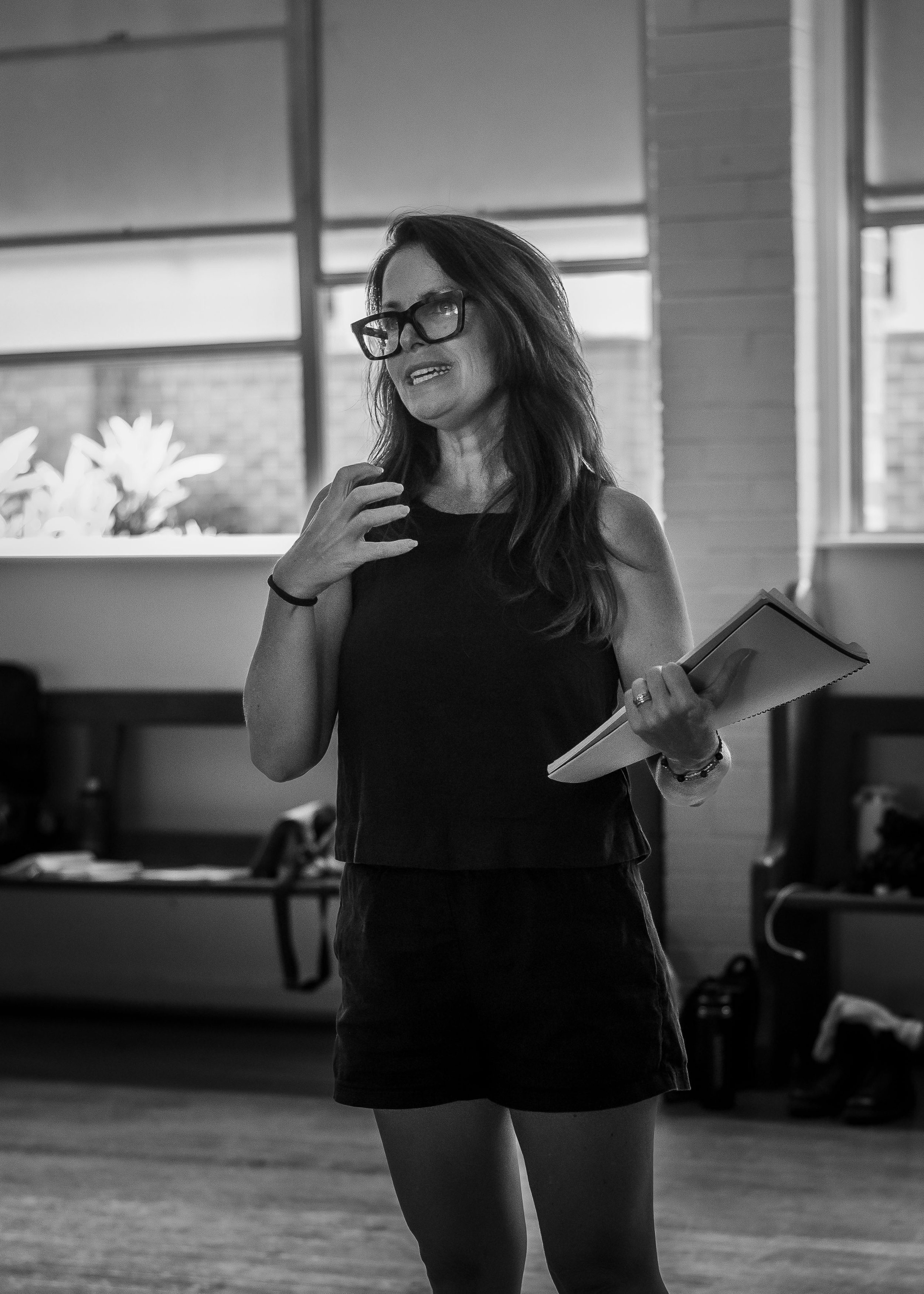 A woman with long hair and glasses holding a notebook, speaking or presenting in a room with large windows and brick walls.