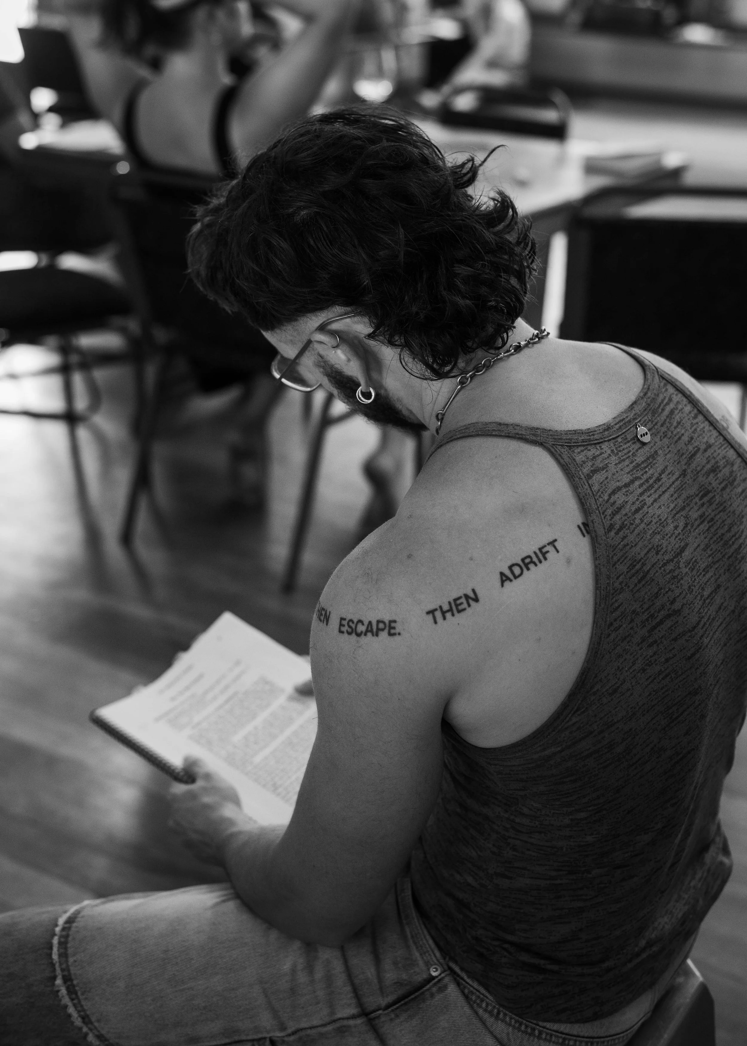 A person with dark curly hair, glasses, earrings, and tattoos sitting on a chair, reading a book in a room with several other people and chairs.