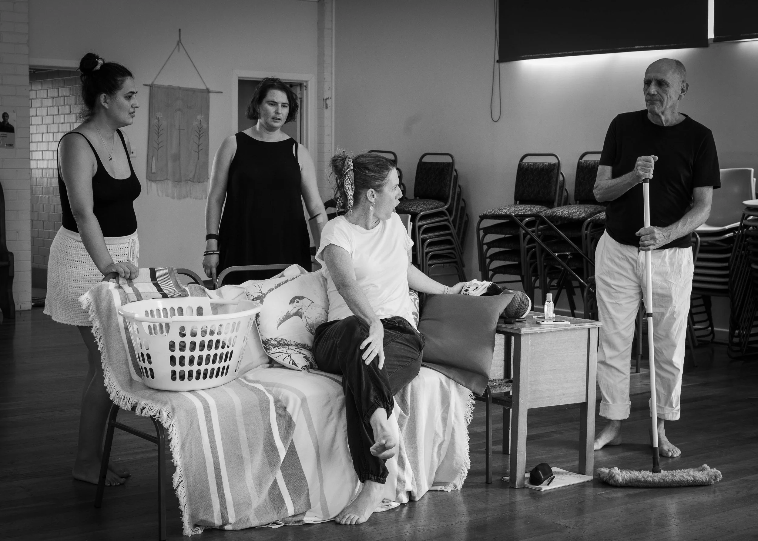 Four women, one sitting on a couch with a pillow, three standing around her, one man with a cane speaking to the women in a casual indoor setting with stacked chairs in the background.