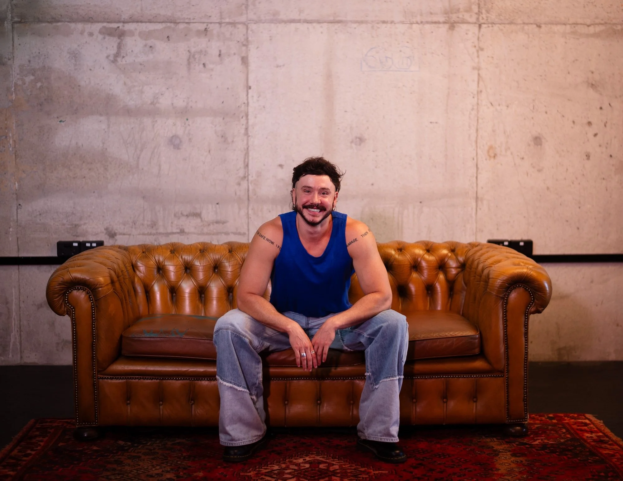 A smiling man with a beard and dark hair, wearing a blue sleeveless shirt and jeans, is sitting on a brown leather tufted sofa in a room with a plain concrete wall background.