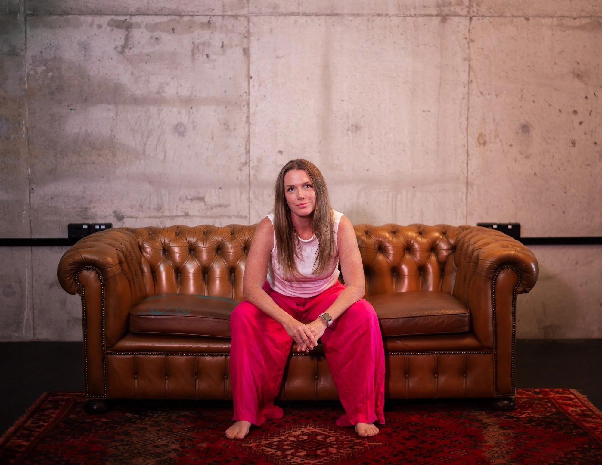 A woman sitting barefoot on a brown leather tufted sofa in front of a plain concrete wall.