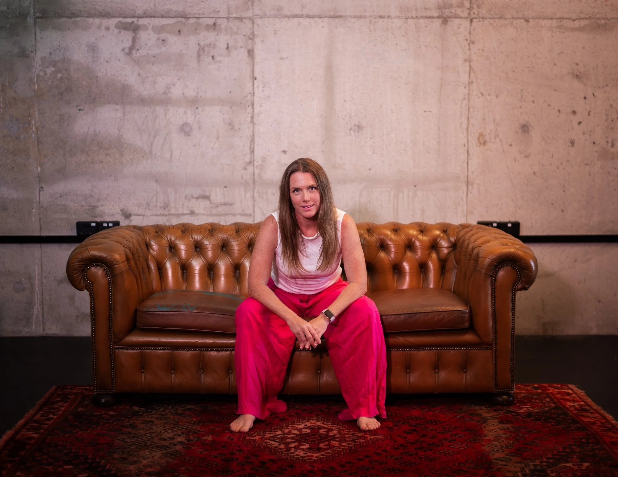 A woman with long brown hair, wearing a white and pink sleeveless shirt and pink pants, sits barefoot on a brown leather tufted sofa in front of a plain concrete wall.