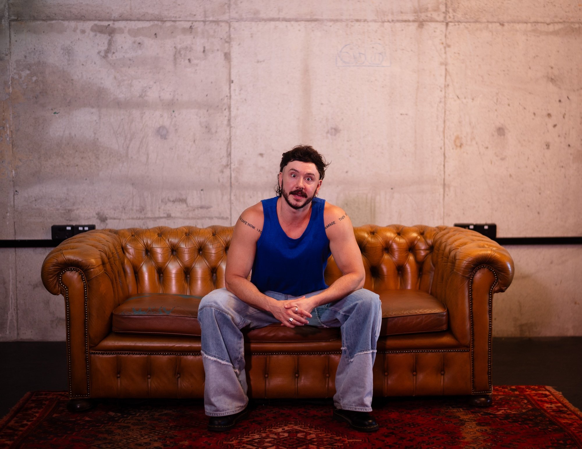 A man with dark hair and a beard wearing a blue tank top and jeans sitting on a brown leather Chesterfield sofa in front of a concrete wall.