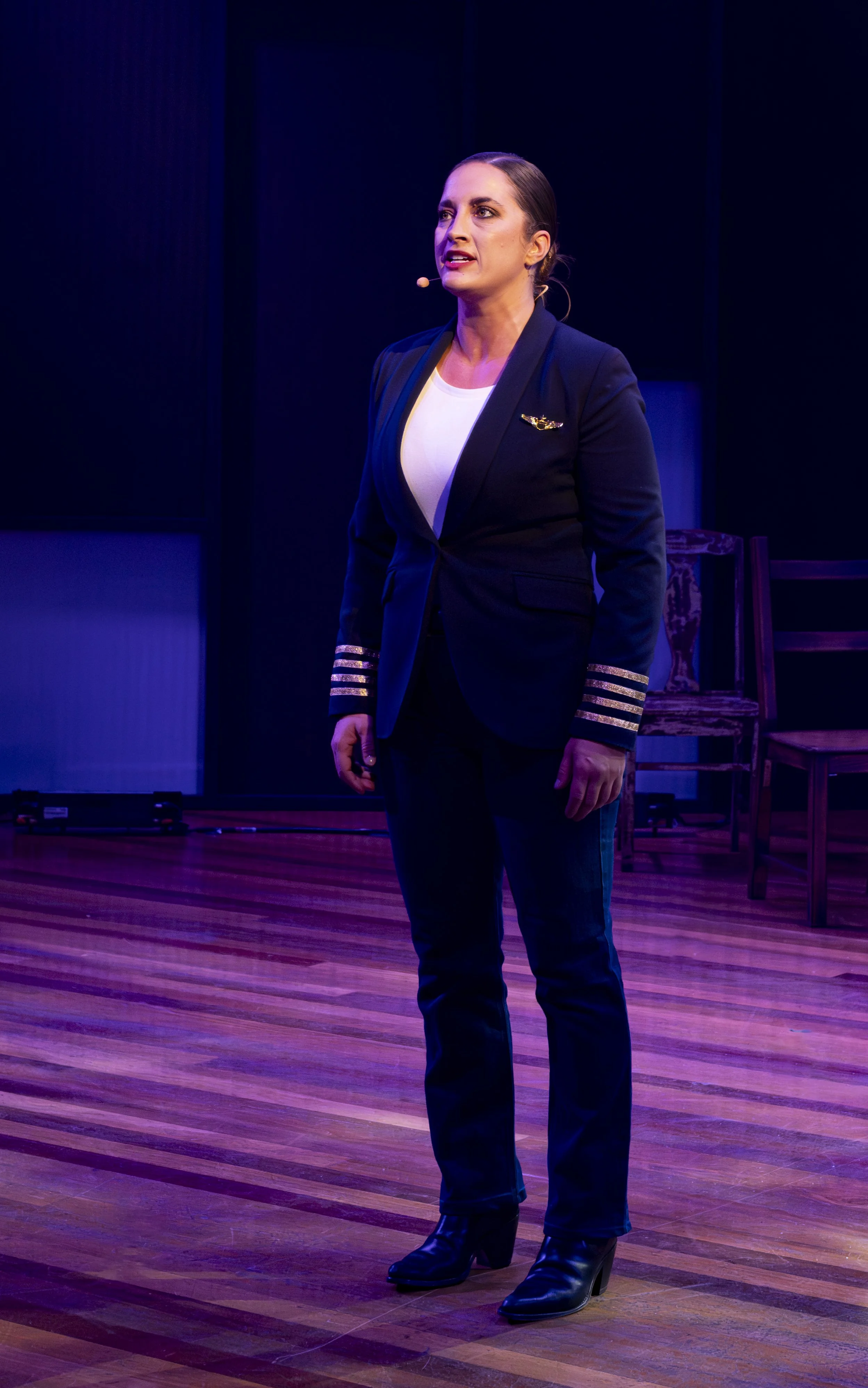 A woman in a navy blazer with gold stripes on the sleeves, standing on a stage with wooden floorboards, speaking into a microphone headset.