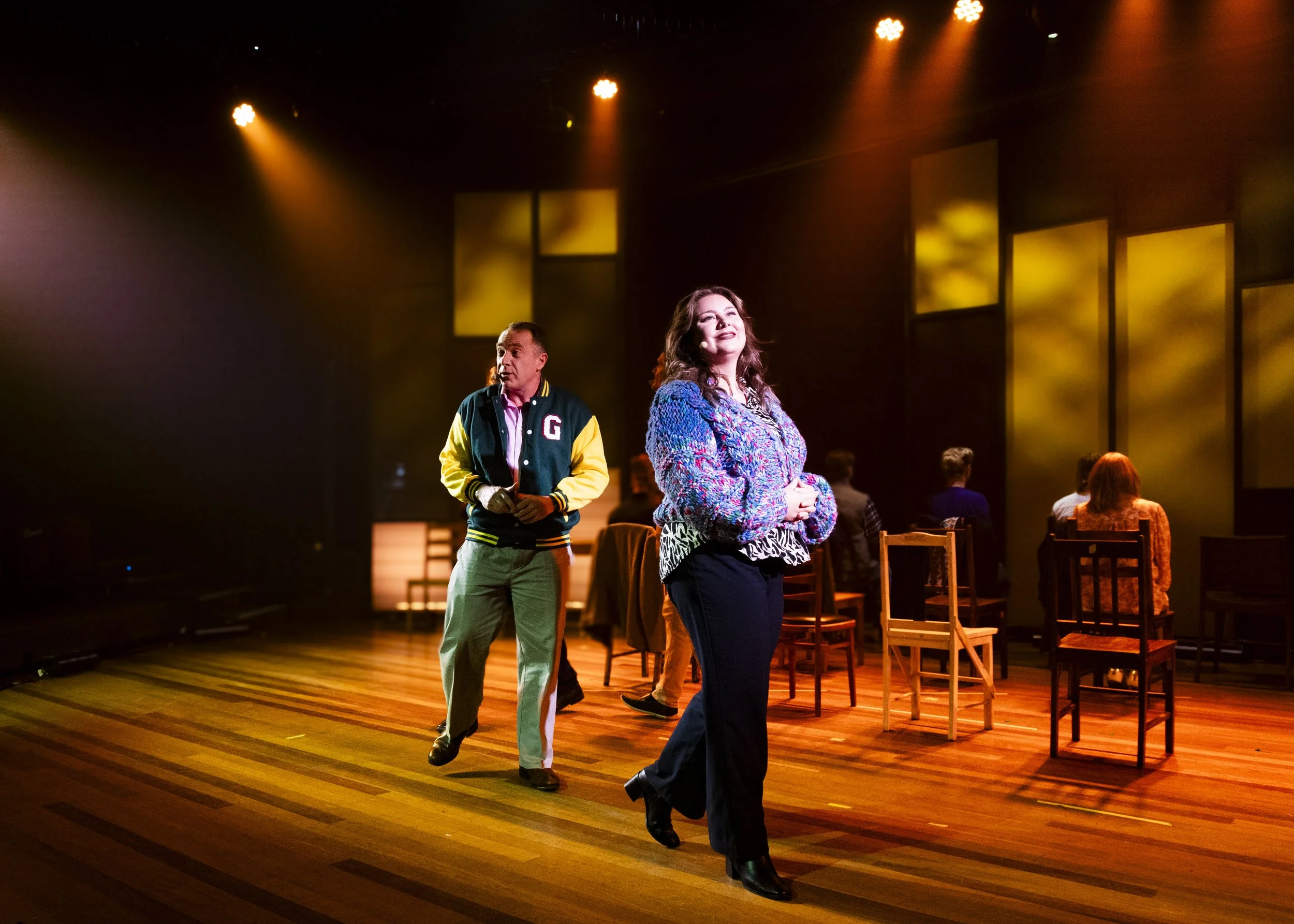Actors performing on stage with warm lighting, audience seated in background, and wooden chairs scattered across the stage.