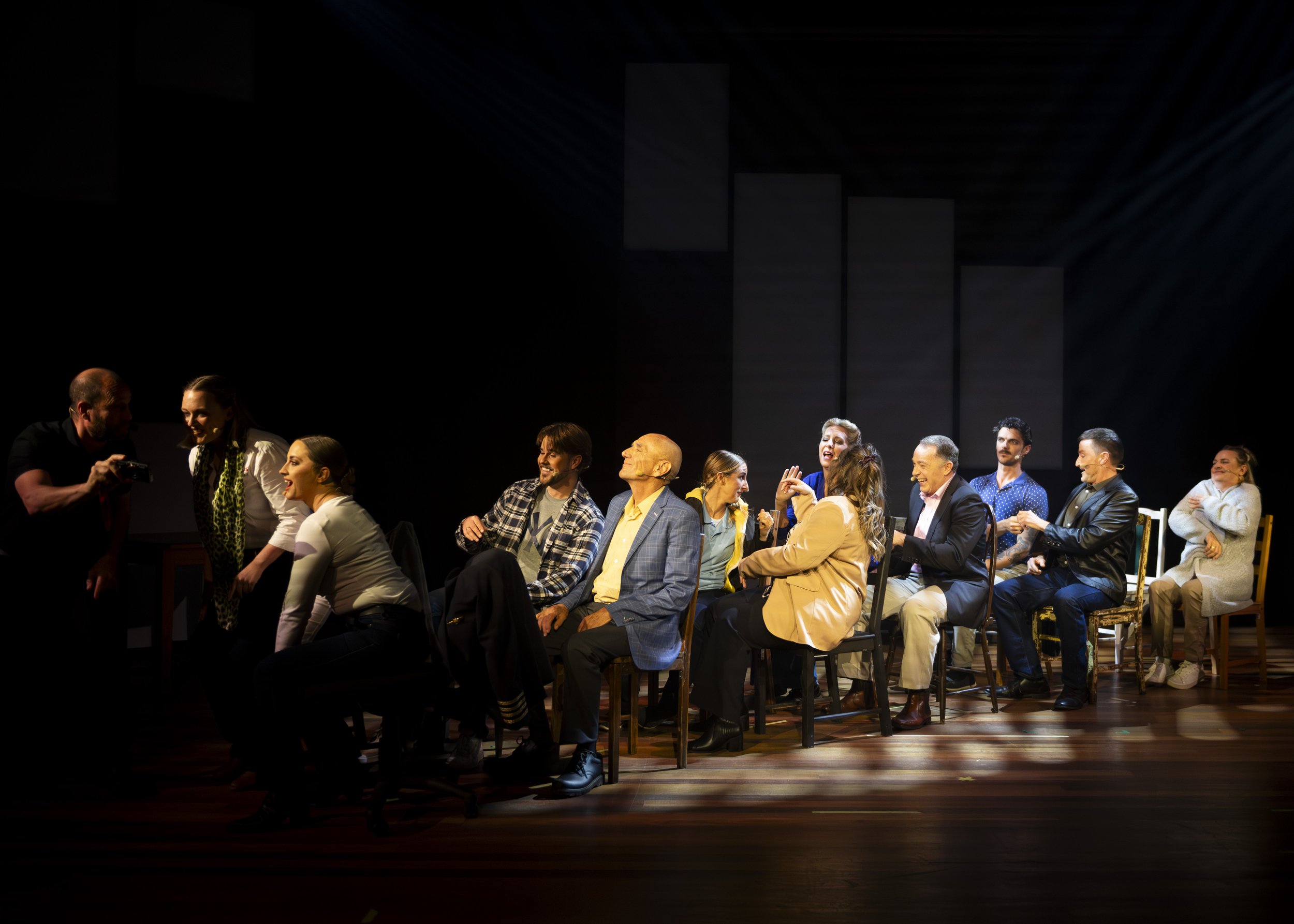 Theater cast on stage with nine actors seated, laughing and talking, while two actors stand to the left, one taking a photo of the group in a darkened theater setting.