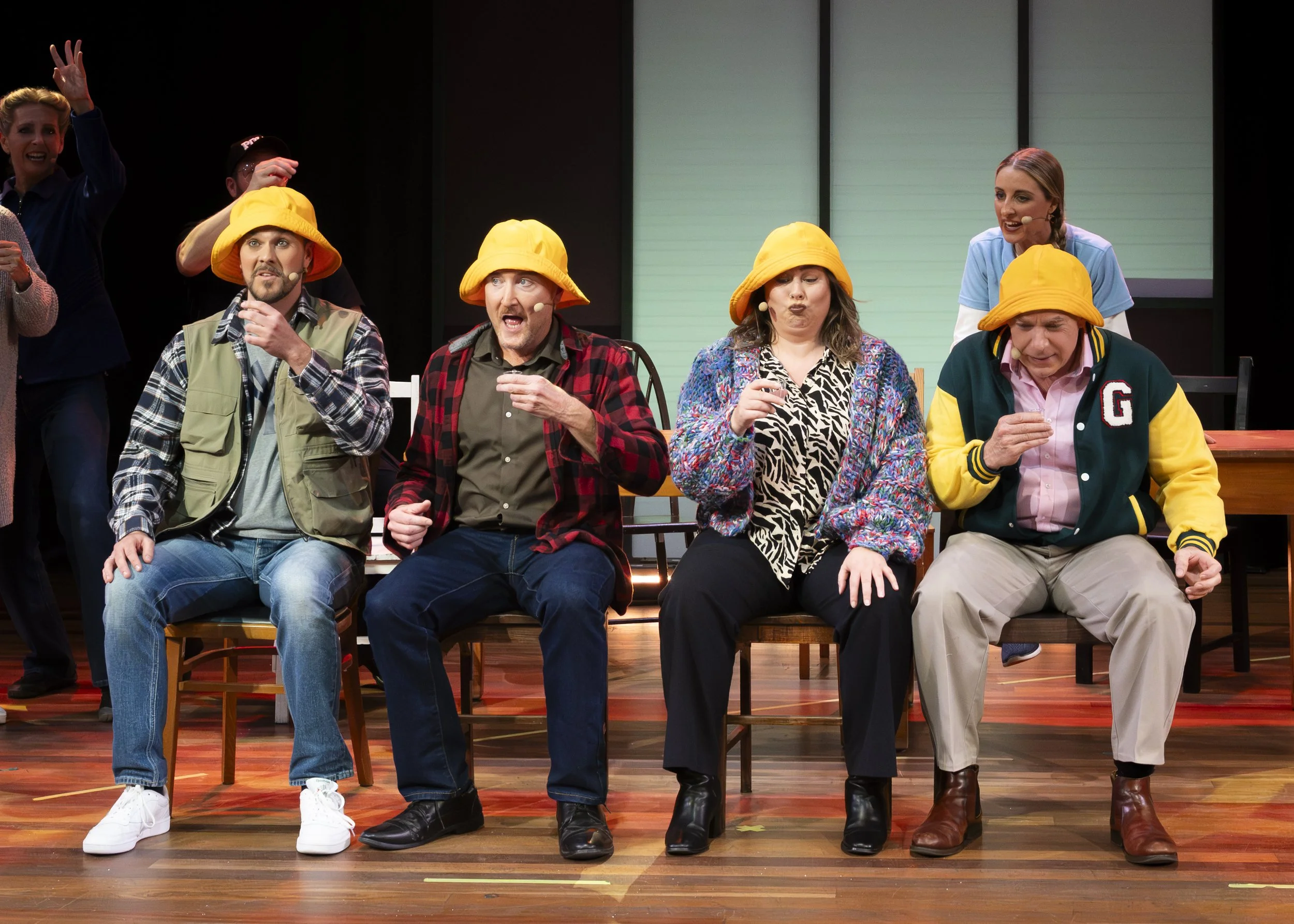 Four performers seated on stage, wearing yellow hats, in theatrical costumes, with their expressions and postures suggesting a comedic scene. Two people stand in the background, one with their hand raised.