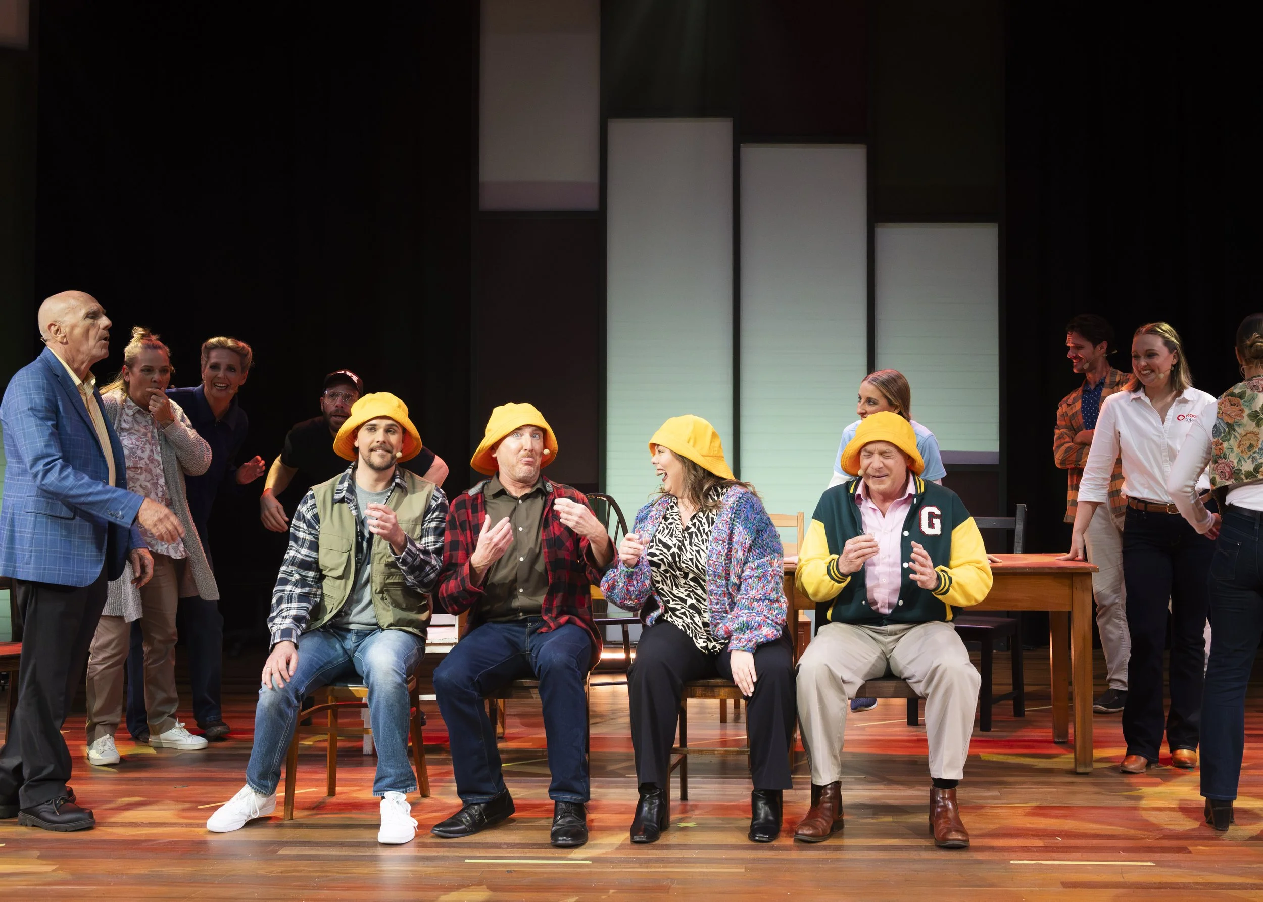 Group of actors on stage during a theater performance, with some wearing yellow hats, seated and standing around a table, with set pieces and lighting in the background.