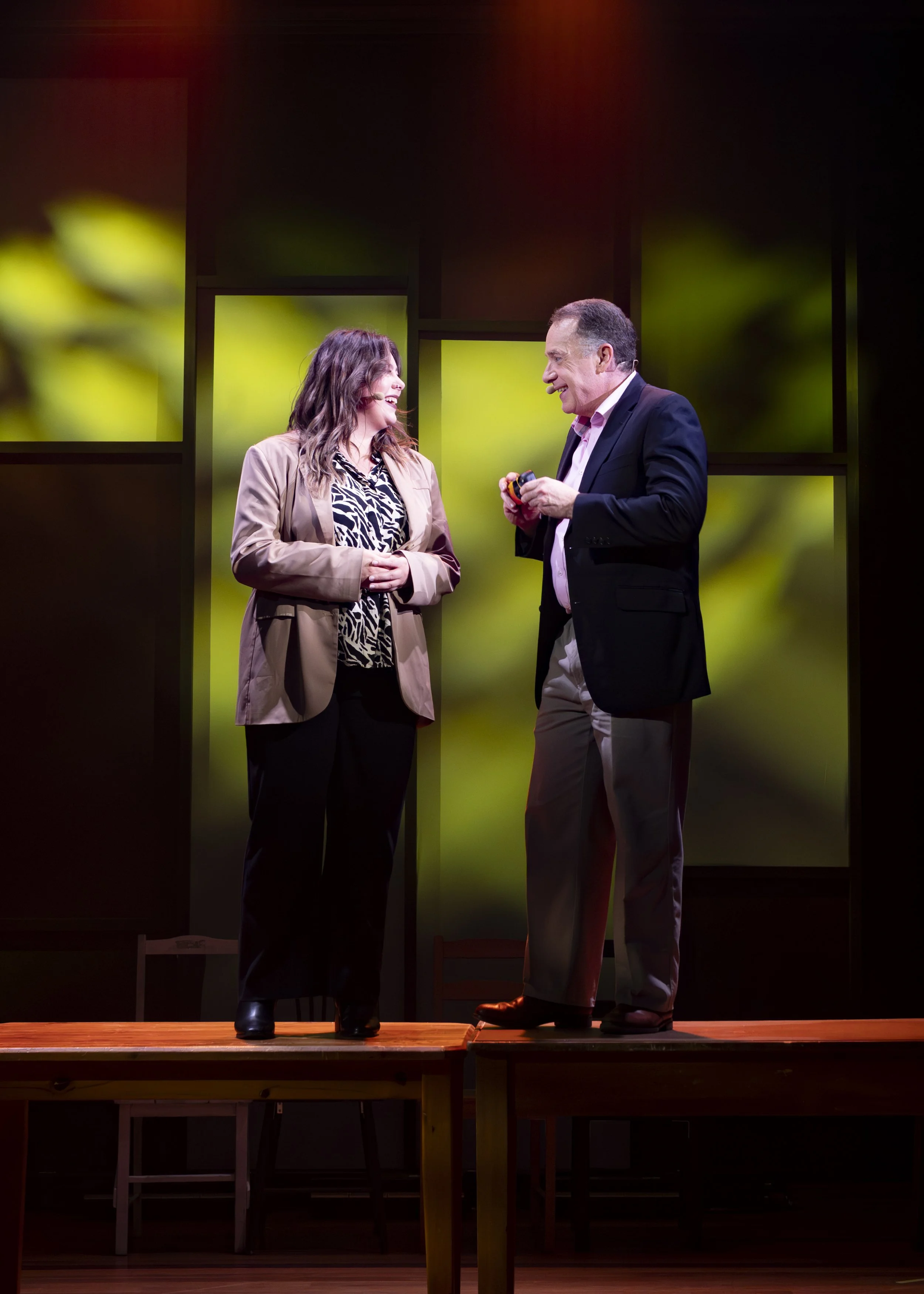 A woman and a man are standing on a stage, engaging in a conversation and smiling at each other. The woman is wearing a beige blazer and black pants, while the man is dressed in a dark blazer and light-colored pants. The background displays a blurred