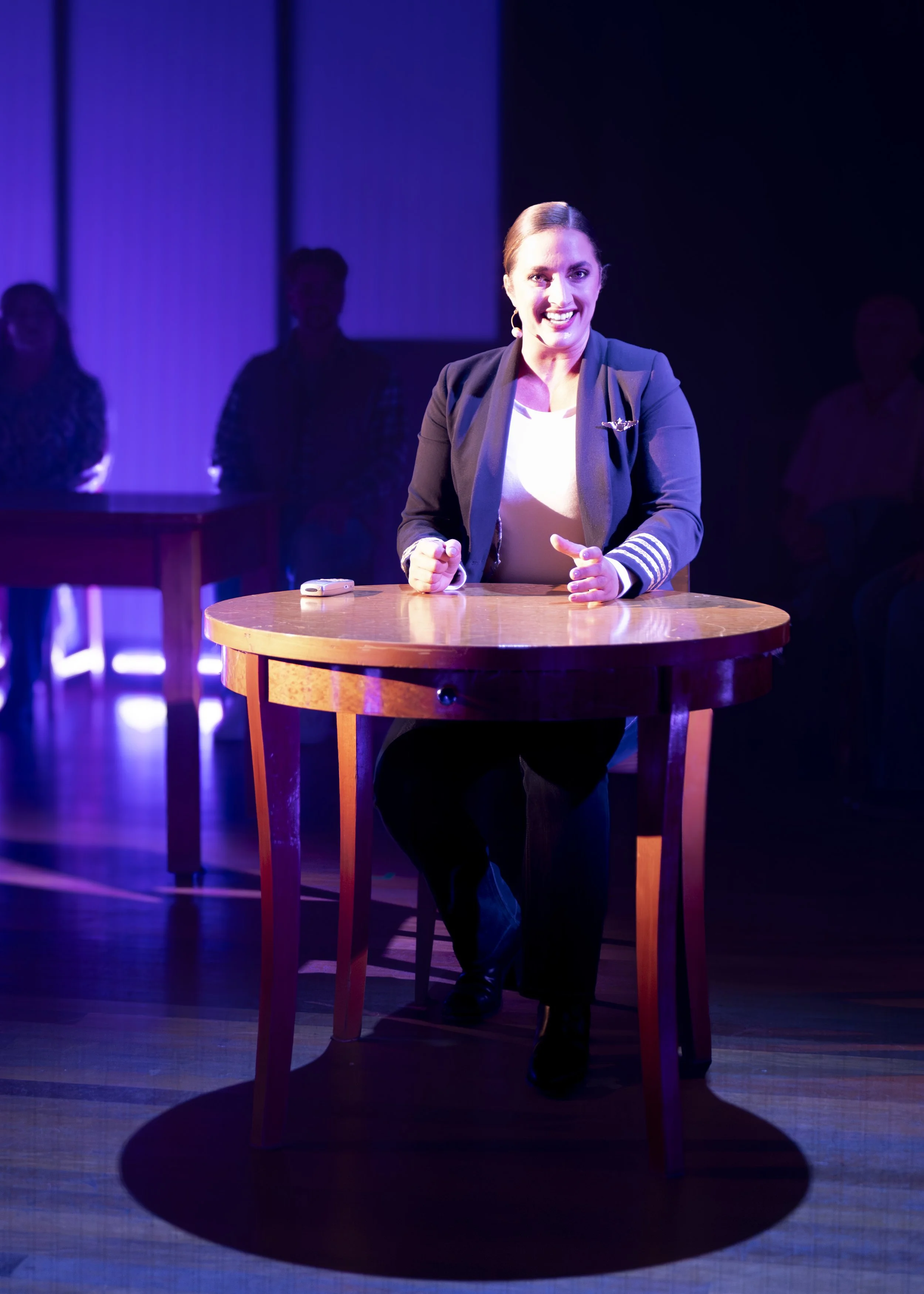 A woman with brown hair, wearing a navy blazer and white shirt, is sitting at a round wooden table on stage, smiling and gesturing with her hands. She is in a spotlight with a dark background and other people sitting in the shadows.