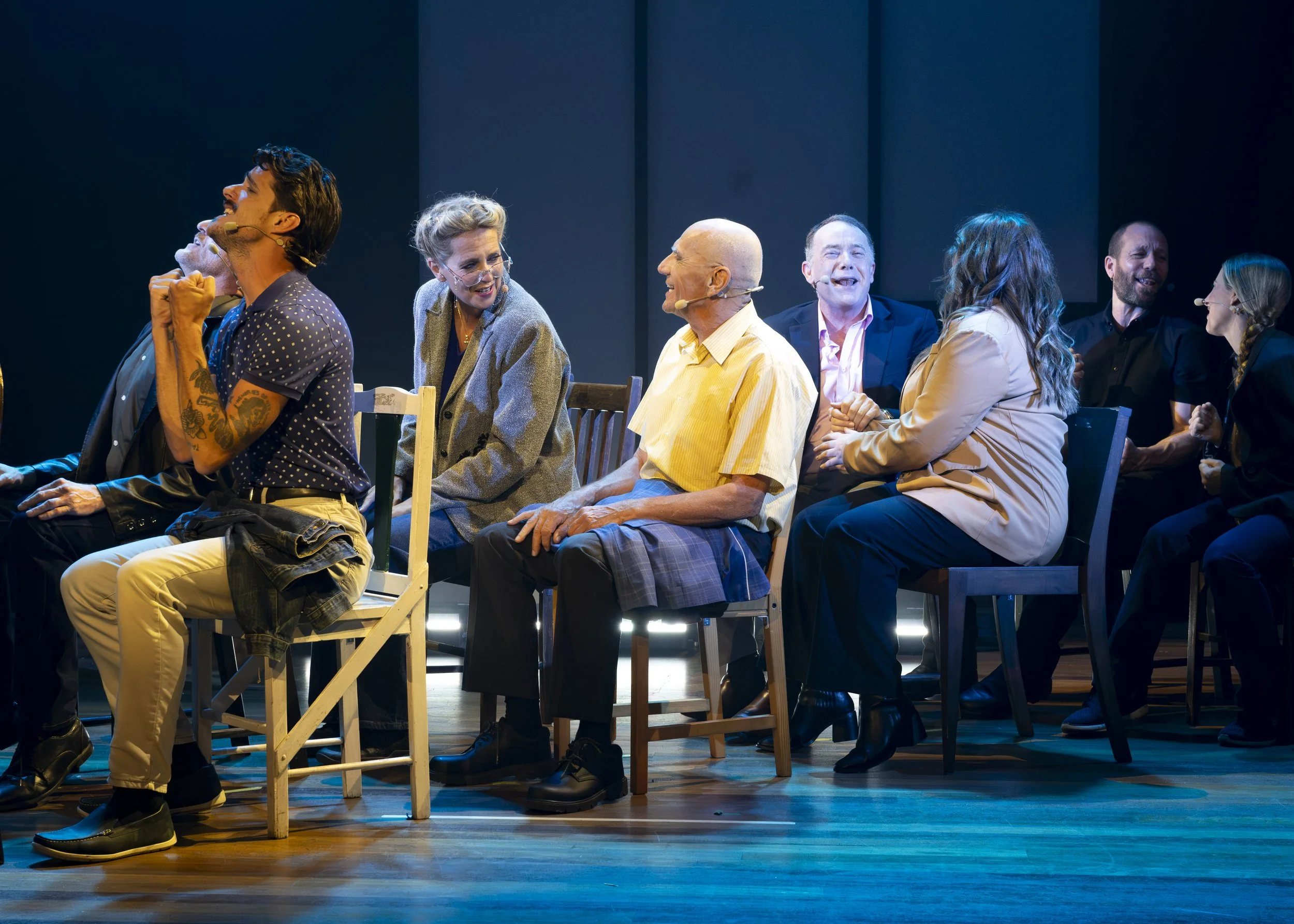 A group of people sitting on chairs on a stage during a theatrical performance, engaging in conversation and laughter.