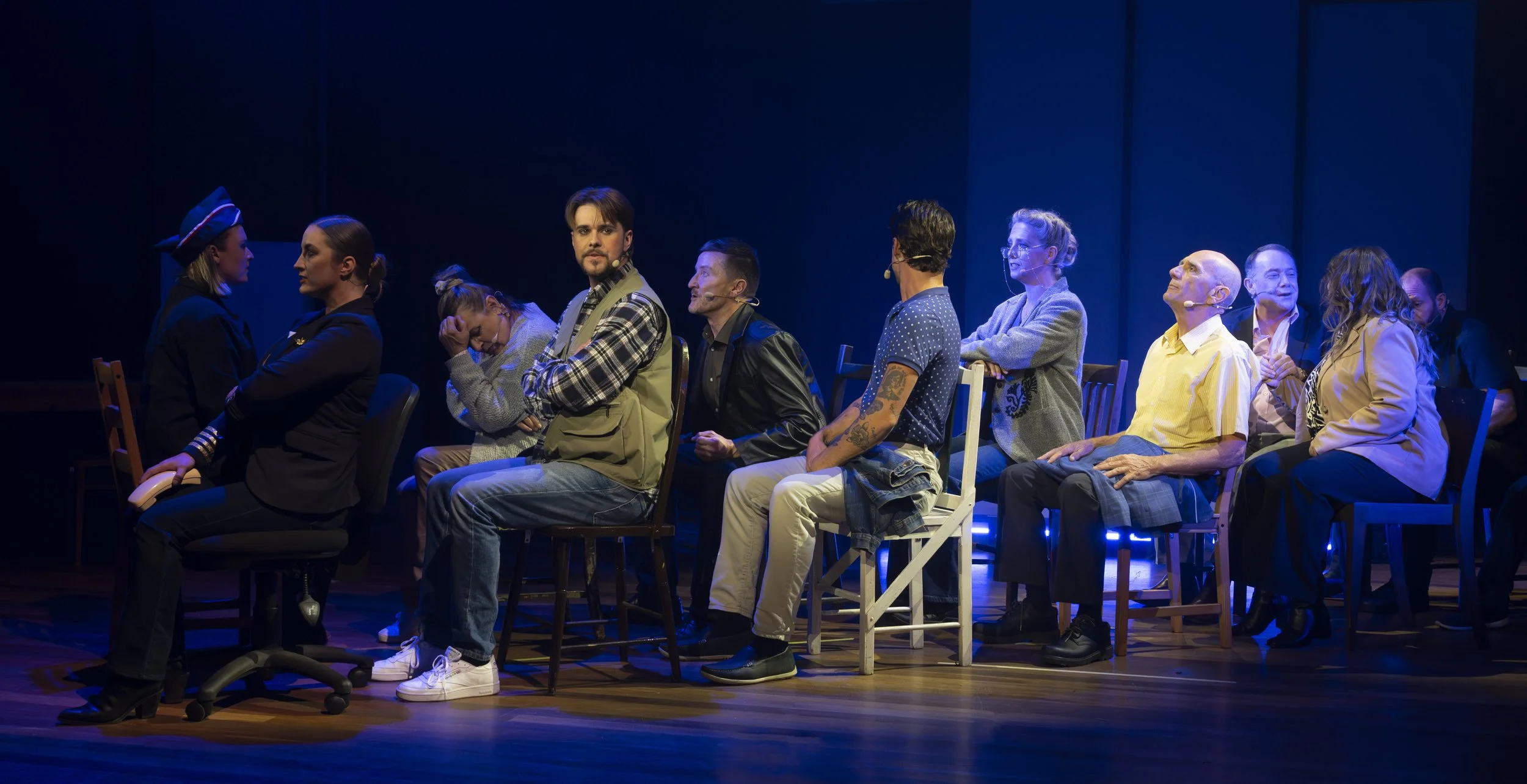Group of people sitting on chairs in a dark theater, some with microphones, engaging in a stage performance or discussion.
