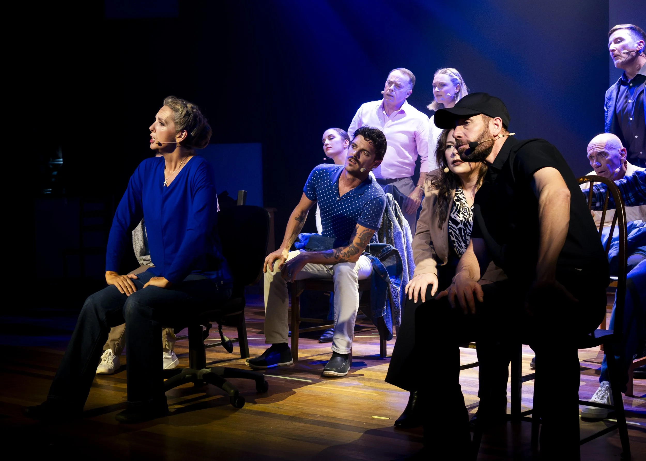A group of diverse people on stage during a theatrical performance, some seated and some standing, with focused and expressive facial expressions, under stage lighting.
